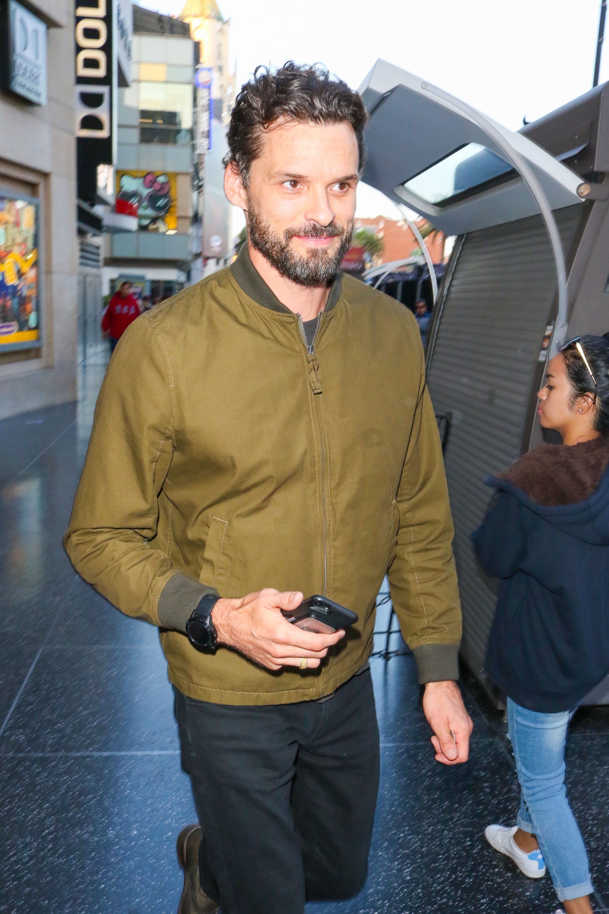 Austin Nichols with a beard in a casual jacket holding a phone, walking down a street with people and shops in the background