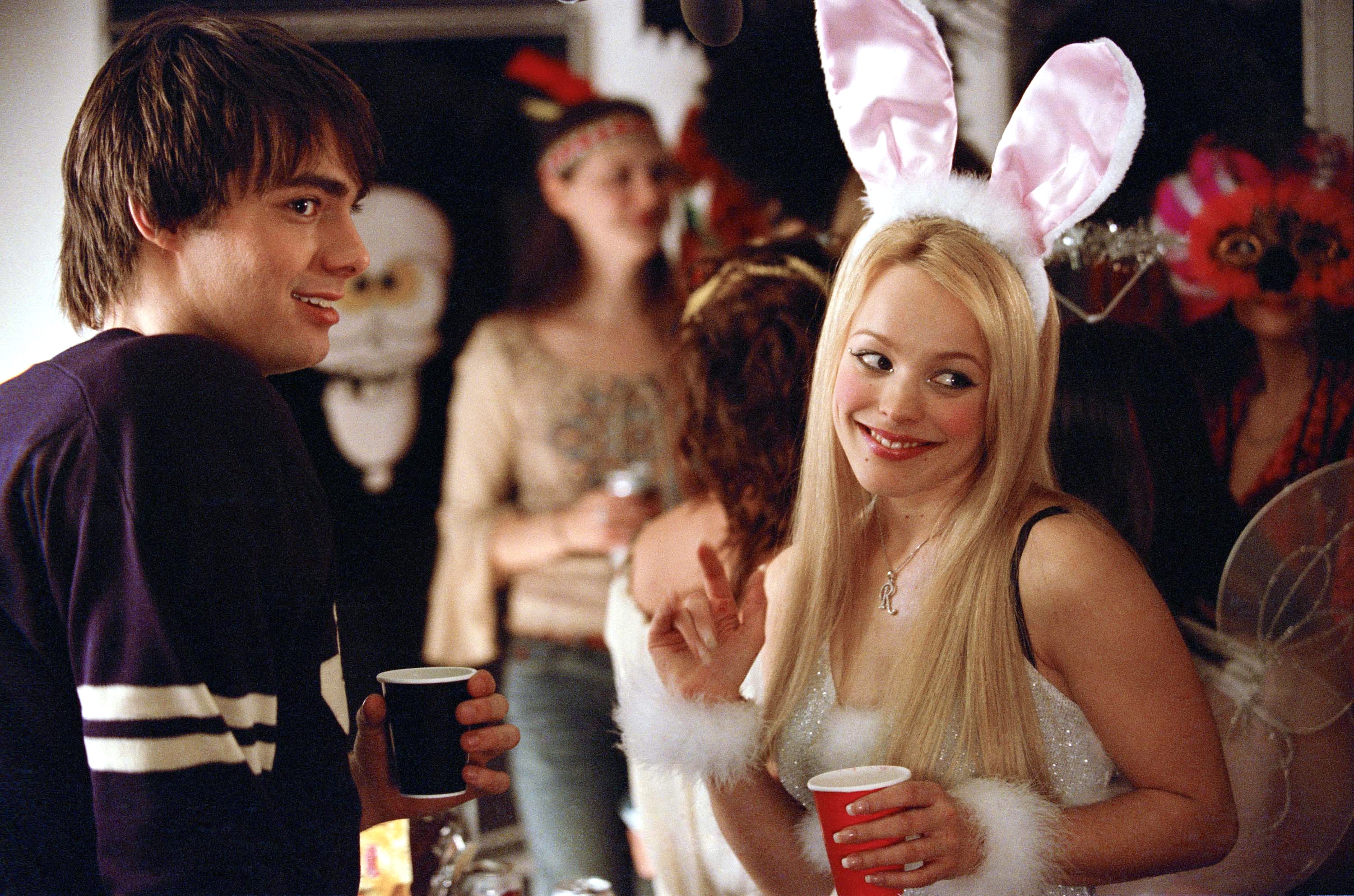 Rachel McAdams as Regina Goerge in bunny ears and a sparkly outfit, smiling and holding a cup. Others in costumes are visible in the background