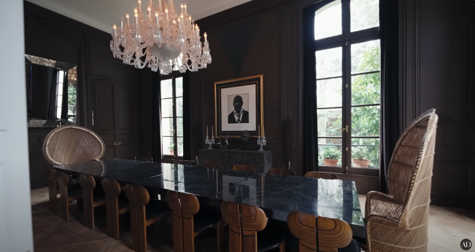 Elegant dining room with a long, dark table, wicker chairs, crystal chandelier, and a framed silhouette portrait above a fireplace