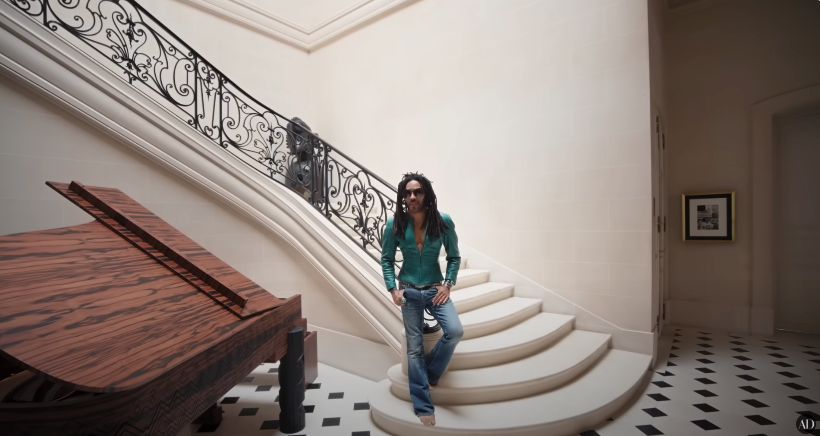 Person with long hair and sunglasses sits on a staircase in a stylish setting with a grand piano and patterned floor, wearing casual jeans and a shirt