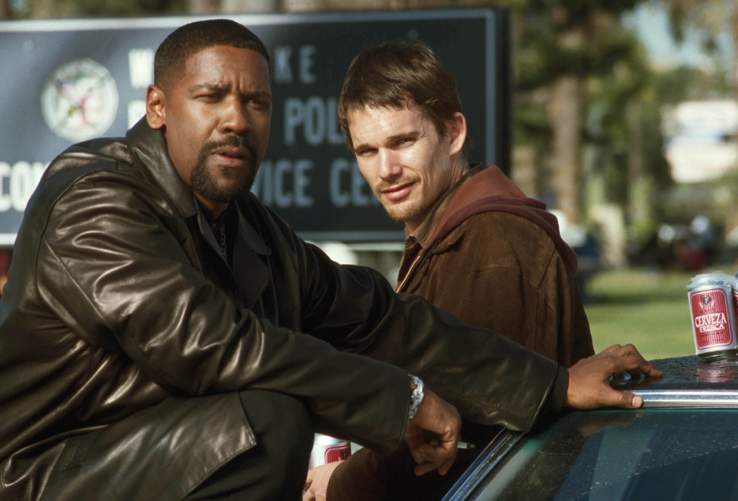 Denzel Washington and Ethan Hawke lean on a car, in a scene from a training day. A police station sign is blurred in the background