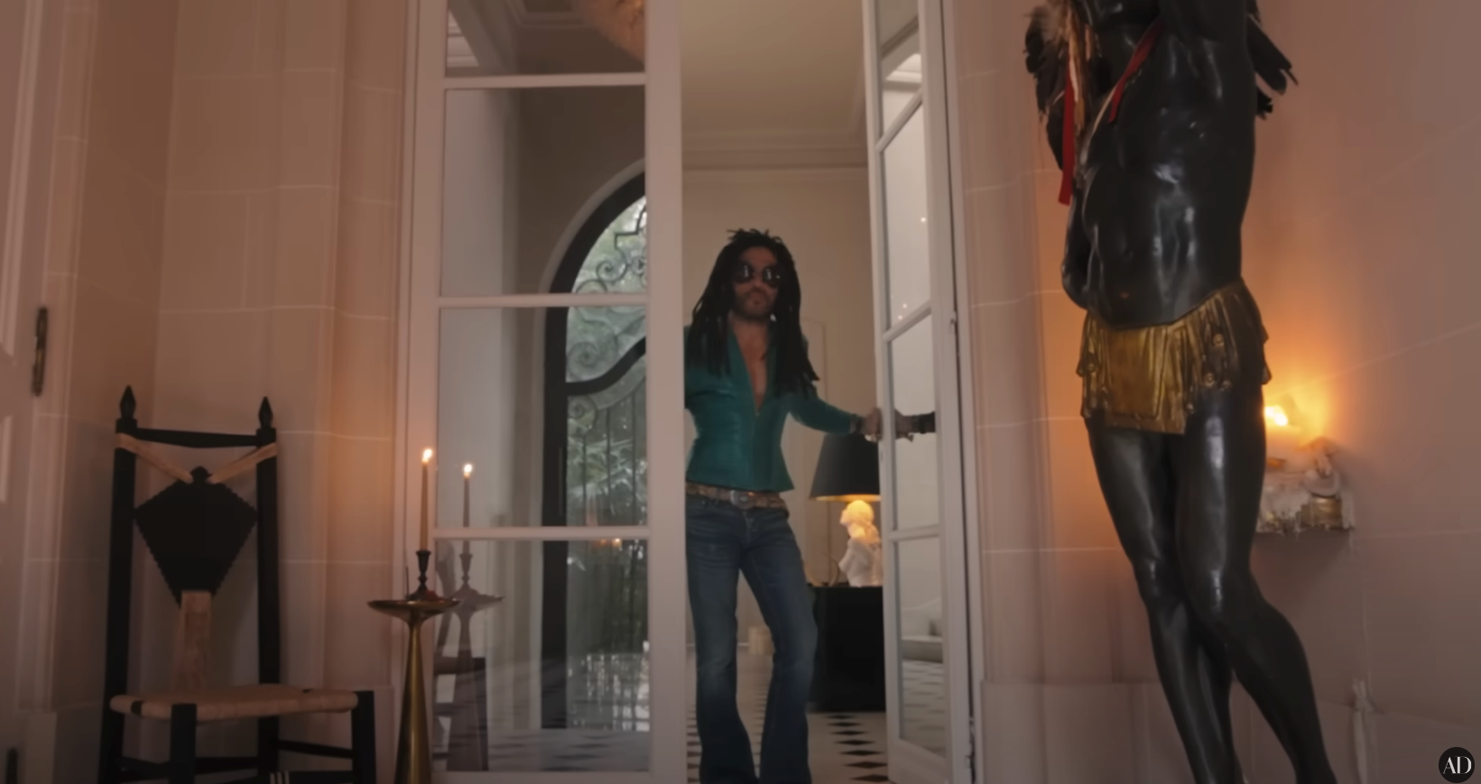 Person in flared jeans and sunglasses entering a room with stylish decor, including a statue and candles