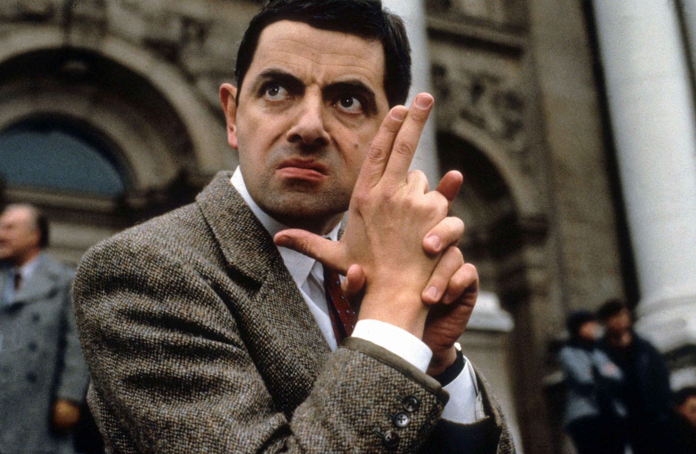 Mr. Bean posing with finger guns, wearing a tweed jacket and white shirt, in front of a building