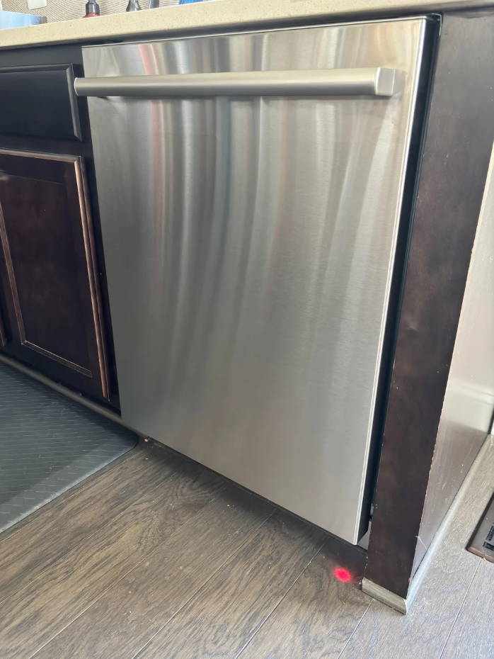 Stainless steel dishwasher with a red light projected onto the wooden floor below, indicating it's running