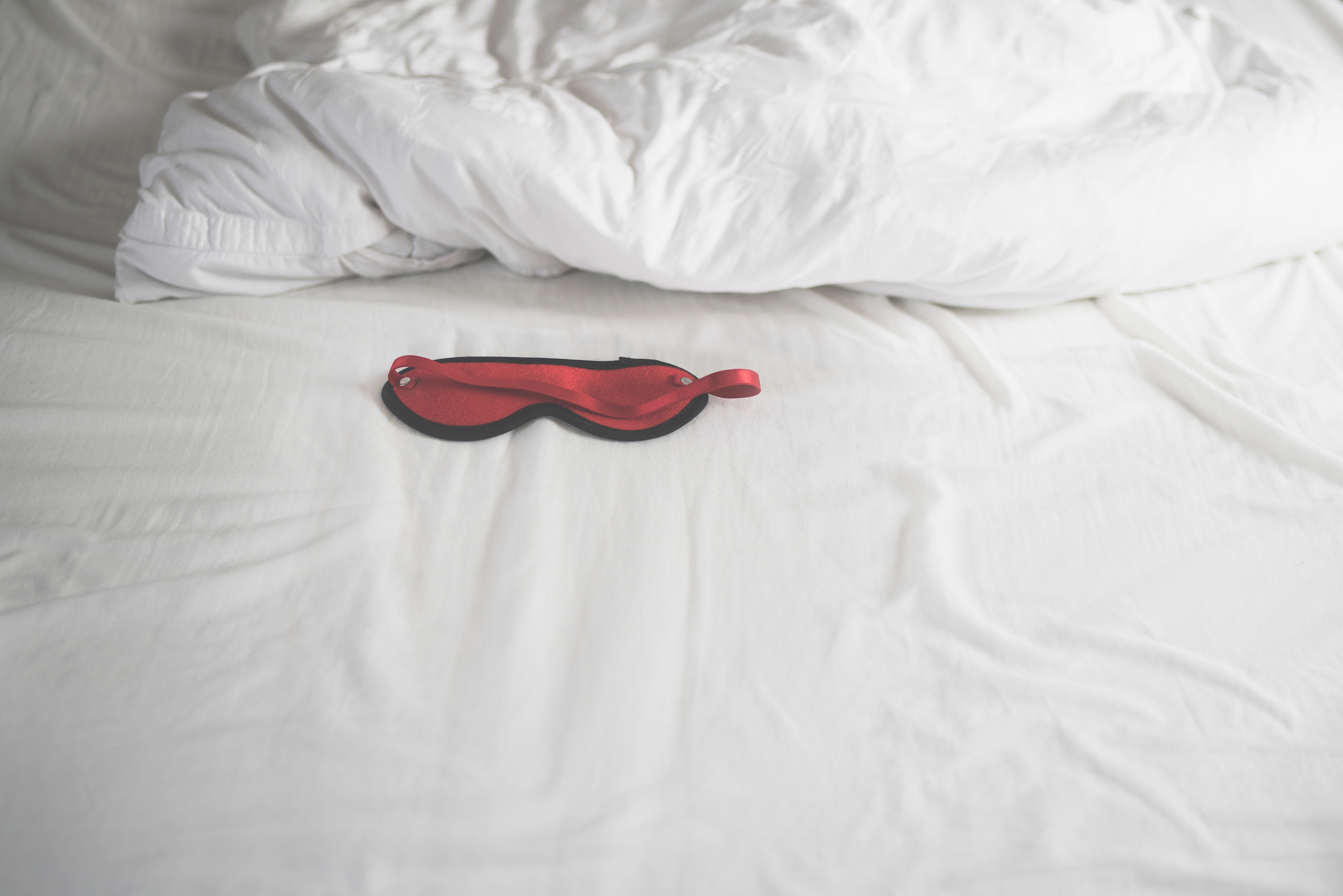 A red satin sleep mask lies on an unmade white bed, implying an intimate or romantic setting