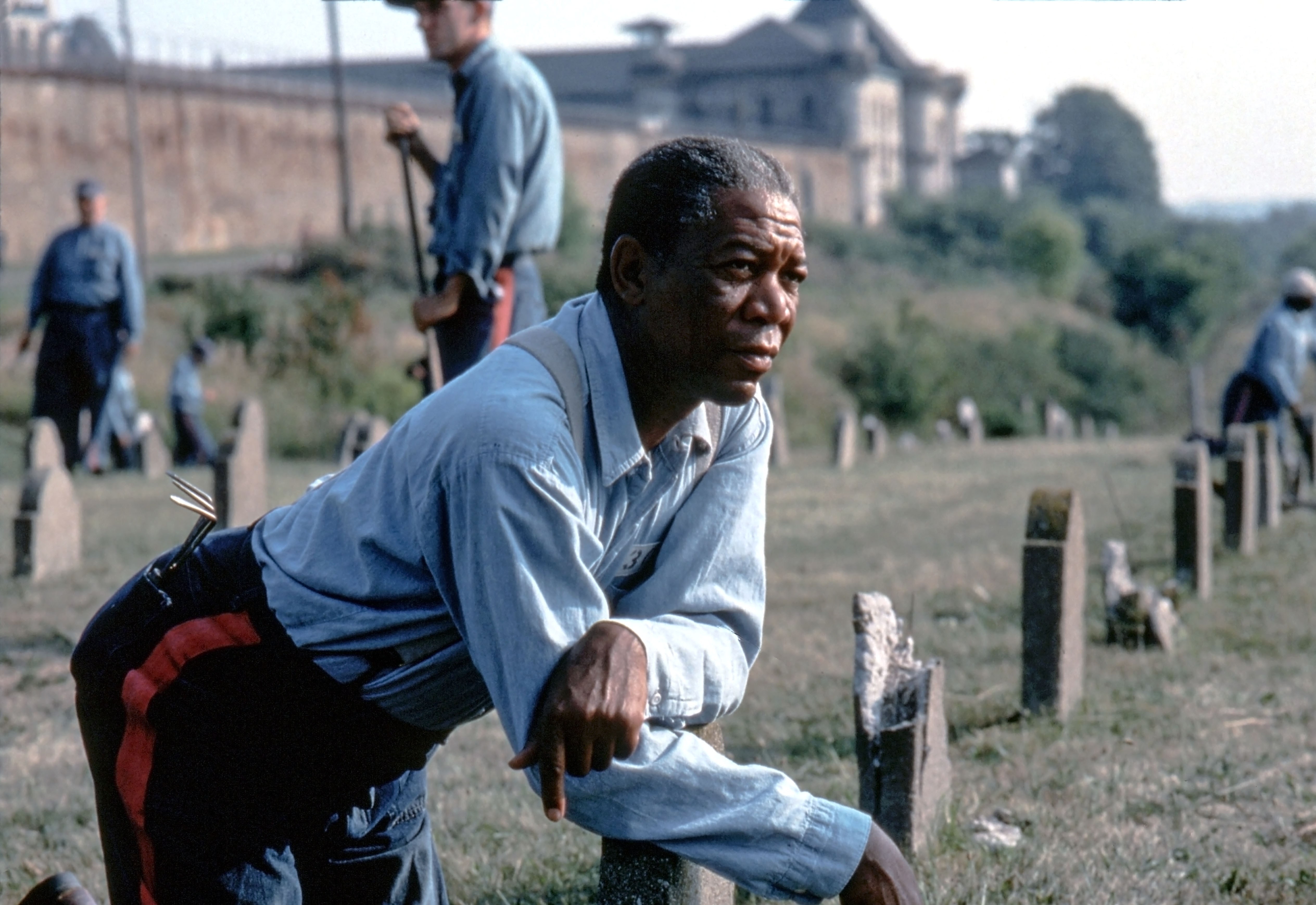 Morgan Freeman as Red leans forward thoughtfully in a grassy area, with others in the background working near gravestones
