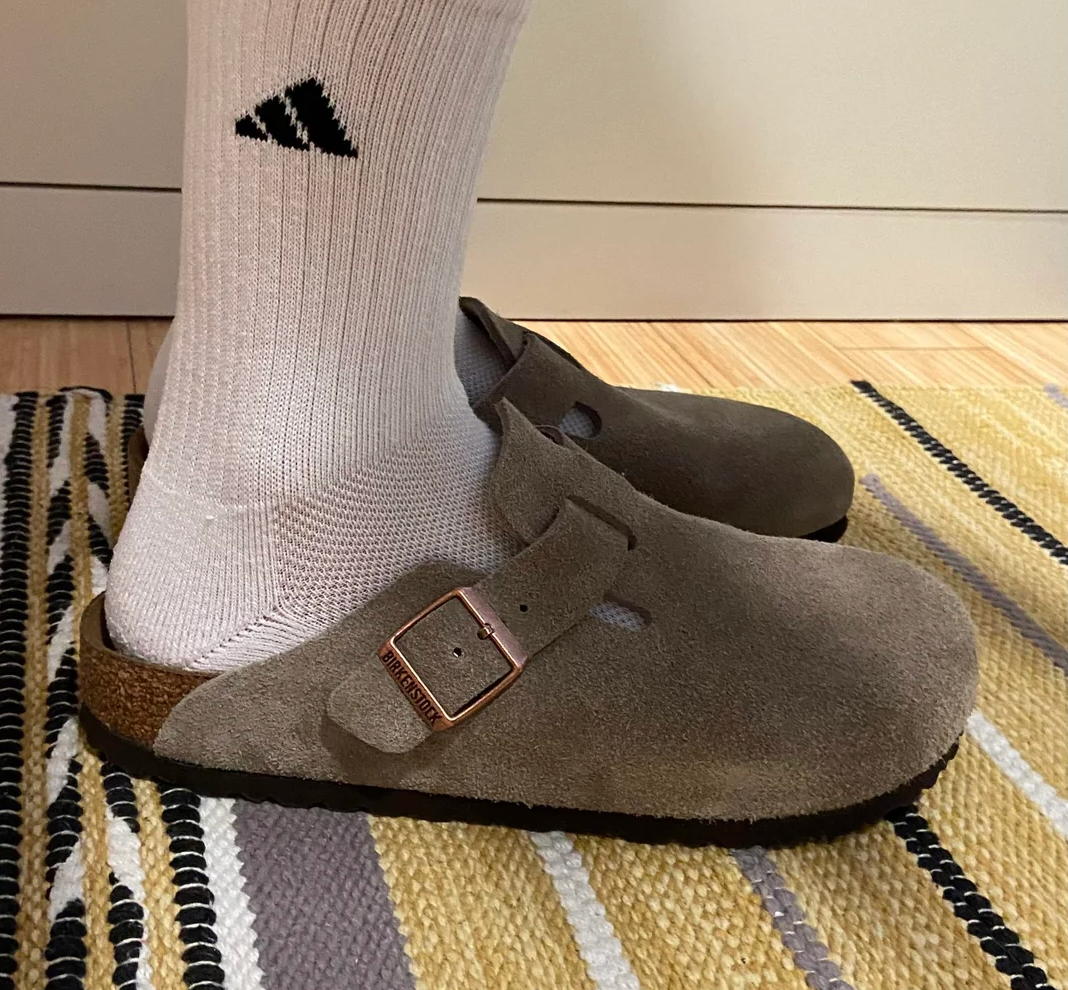 Person wearing white socks and brown suede slip-on clogs, standing on a striped rug