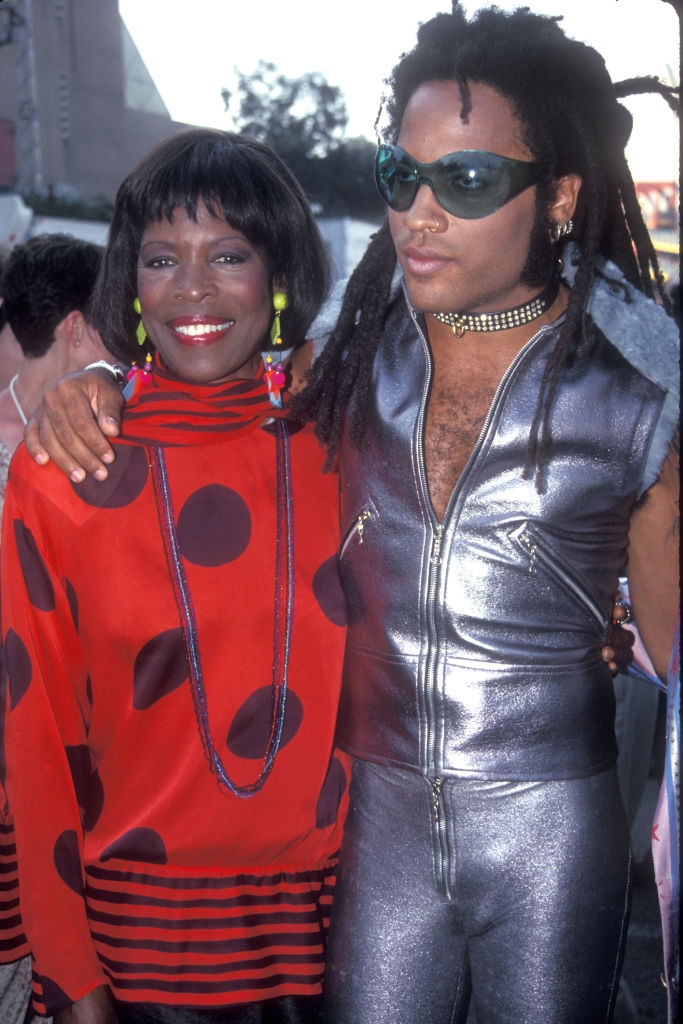 Two people posing together at an event; one in a polka-dot top and the other in a shiny, metallic outfit with sunglasses and a choker