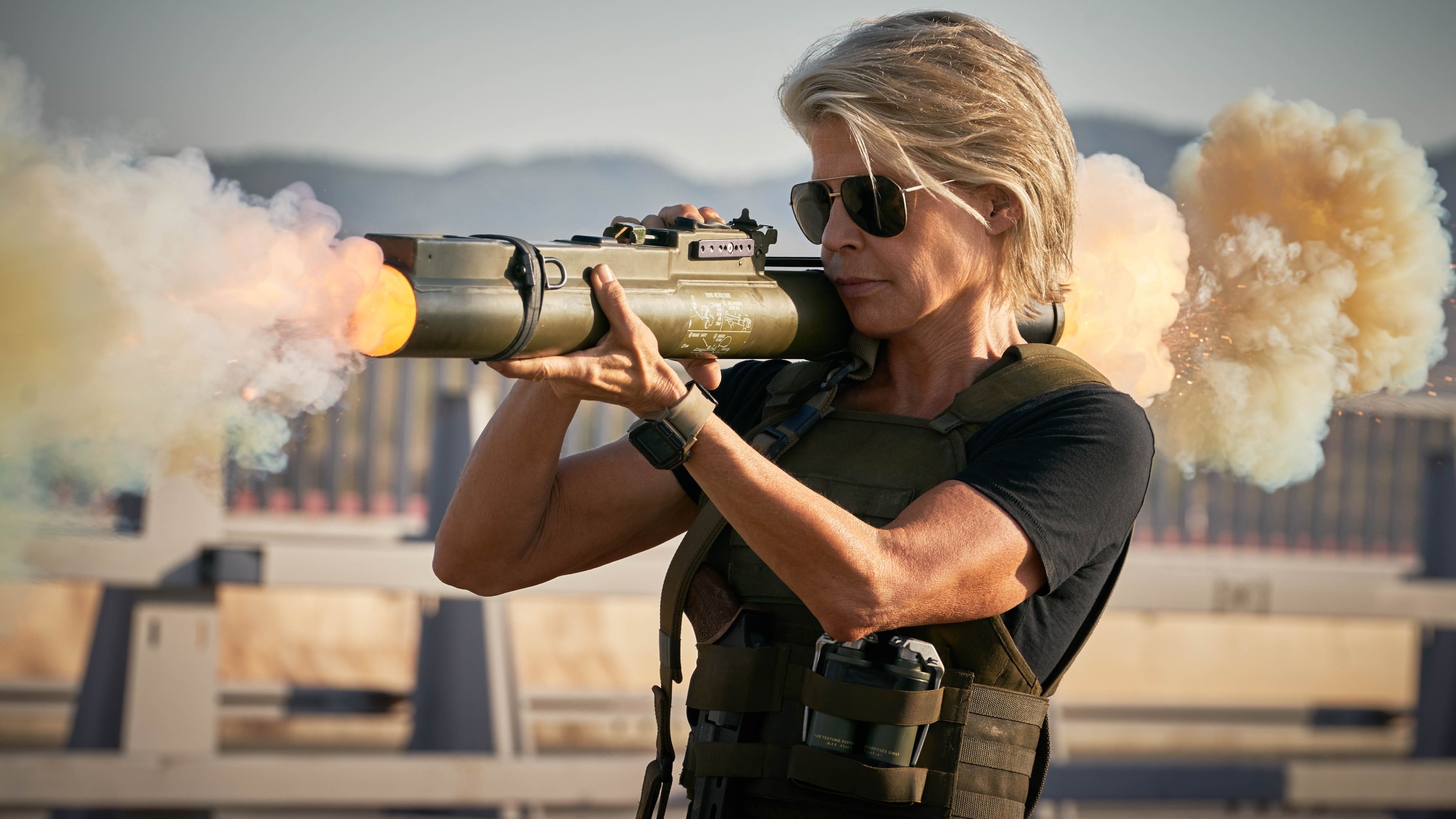 Linda as Sara Conner aiming a rocket launcher outdoors, wearing tactical gear and sunglasses, with smoke emanating from the weapon