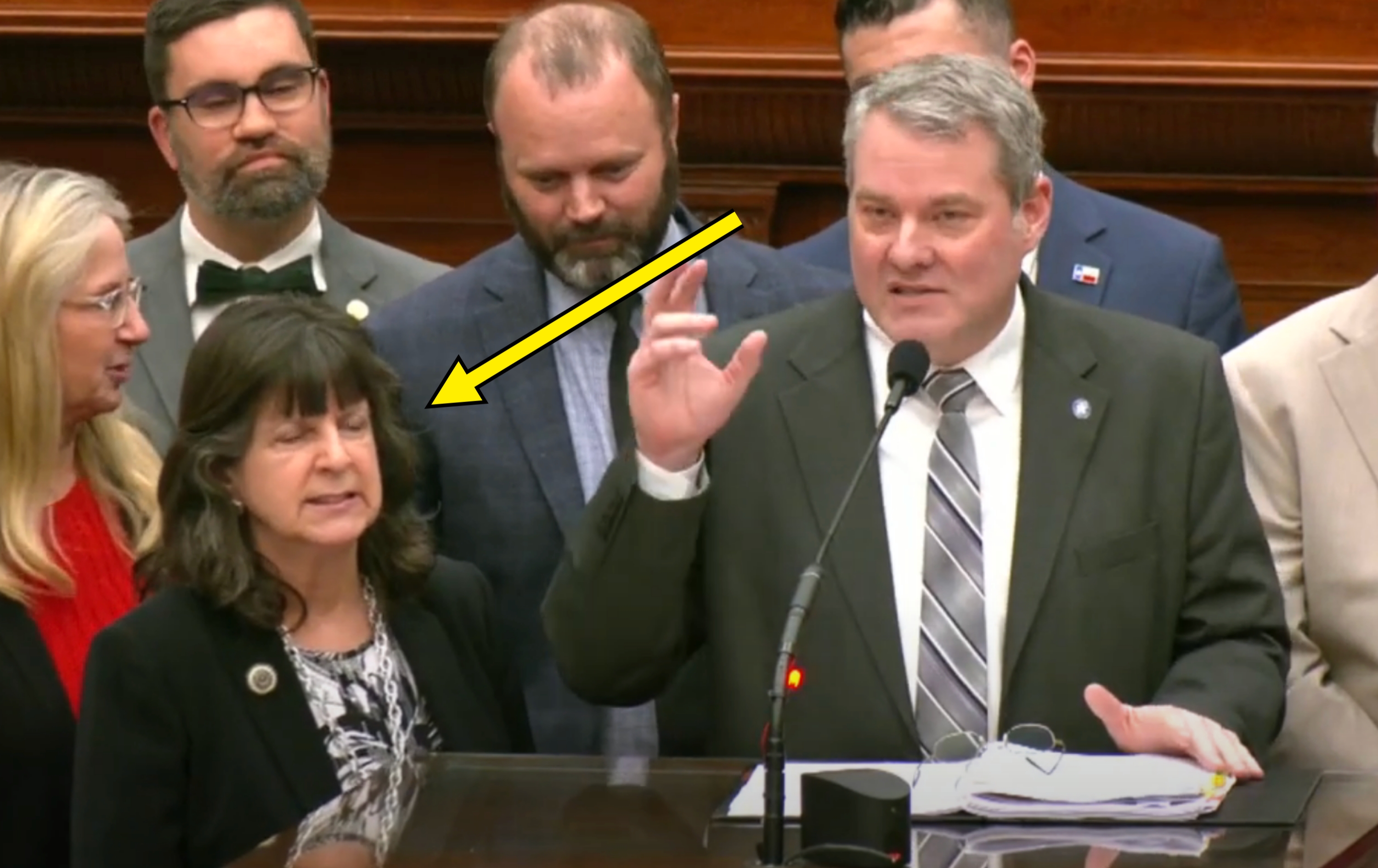 Group of people standing at a podium in a formal setting, one man speaking, others listening attentively