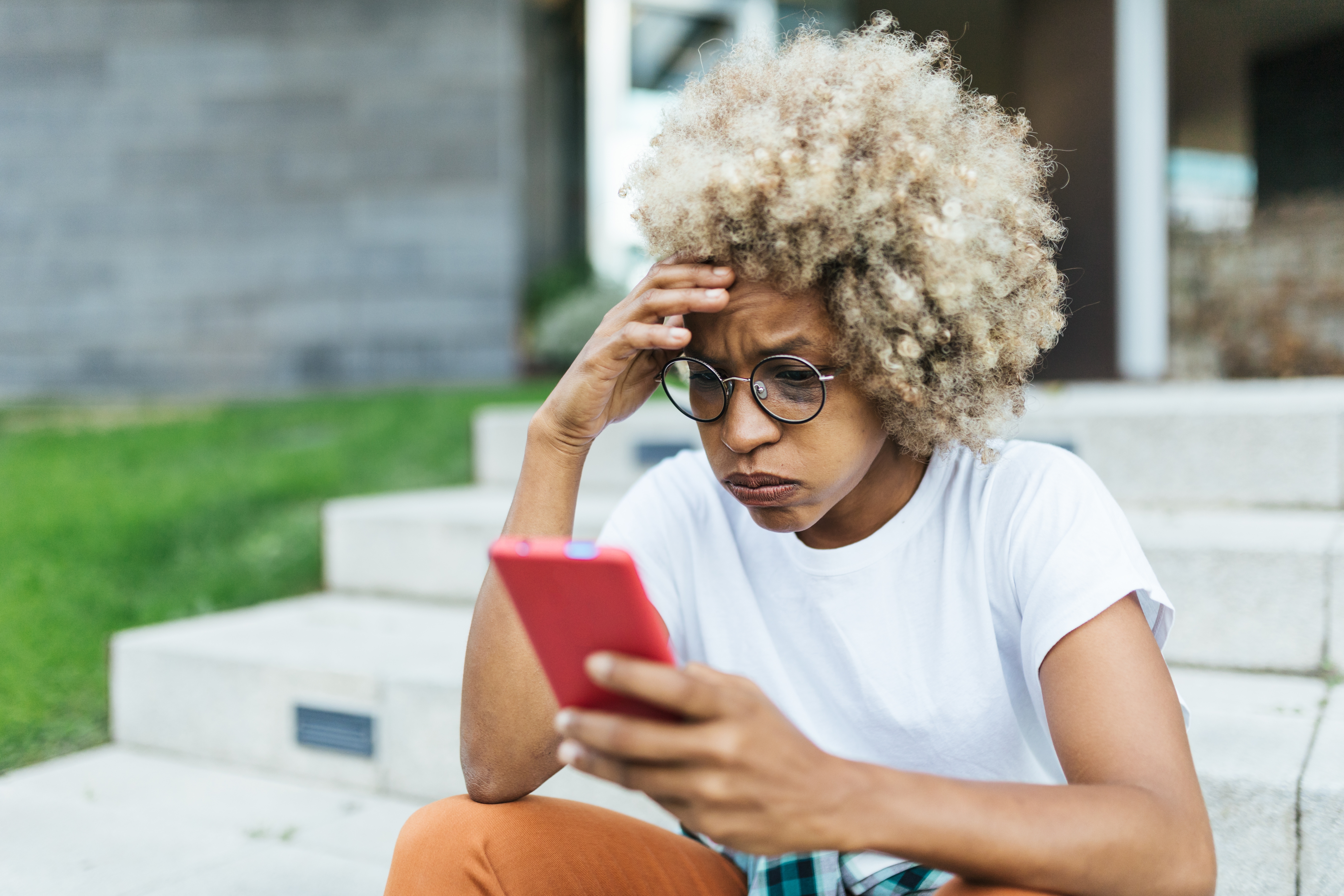 Person with glasses and curly hair sits on steps, looking intently at their smartphone, appearing focused or concerned
