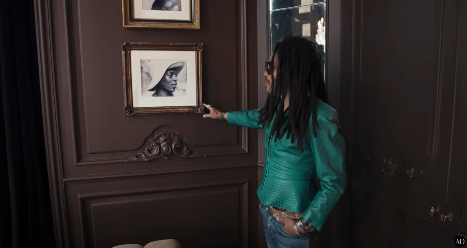 Person in stylish green jacket with long hair points at framed black-and-white photograph on a wall. Interior decor elements are visible