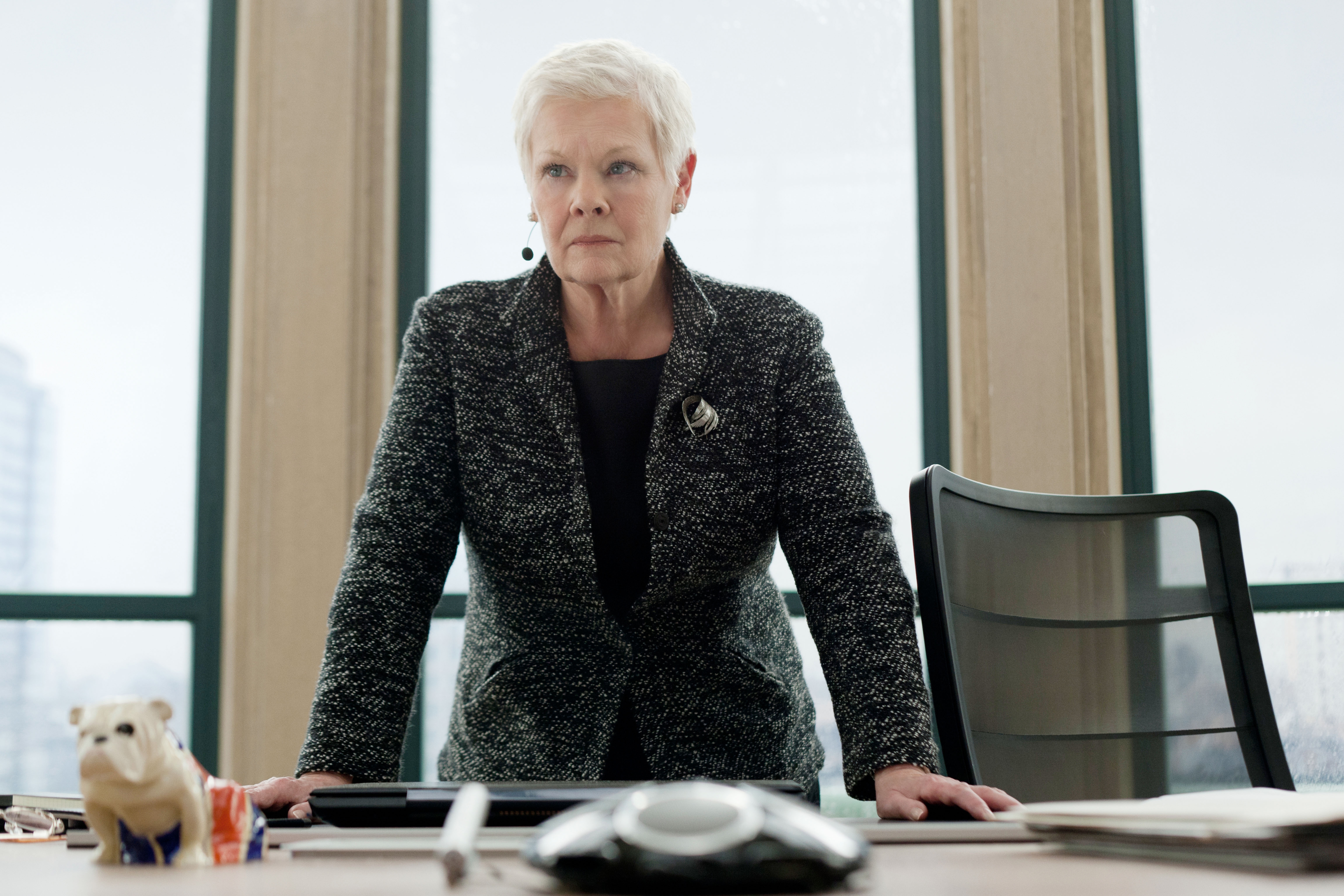 Judi Dench as M in an office leans on a desk, wearing a tweed blazer and brooch, with a small polar bear figure nearby