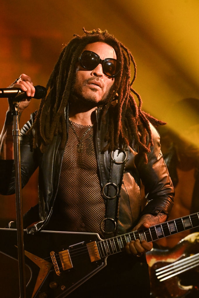 Musician with dreadlocks and sunglasses performs on stage, wearing a leather jacket and mesh shirt, playing an electric guitar