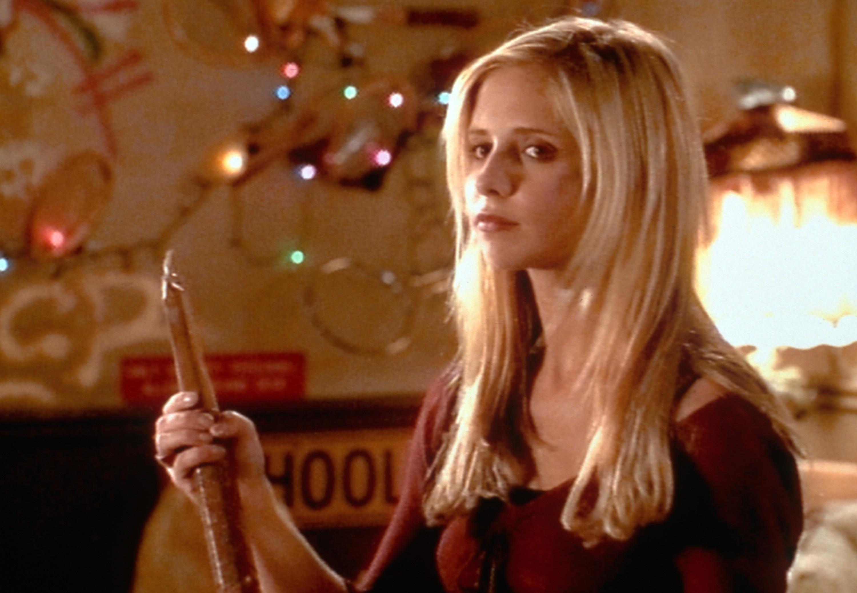 Sarah Michelle Geller as Buffy holds a wooden stake, standing in a room with soft lighting and colorful lights in the background