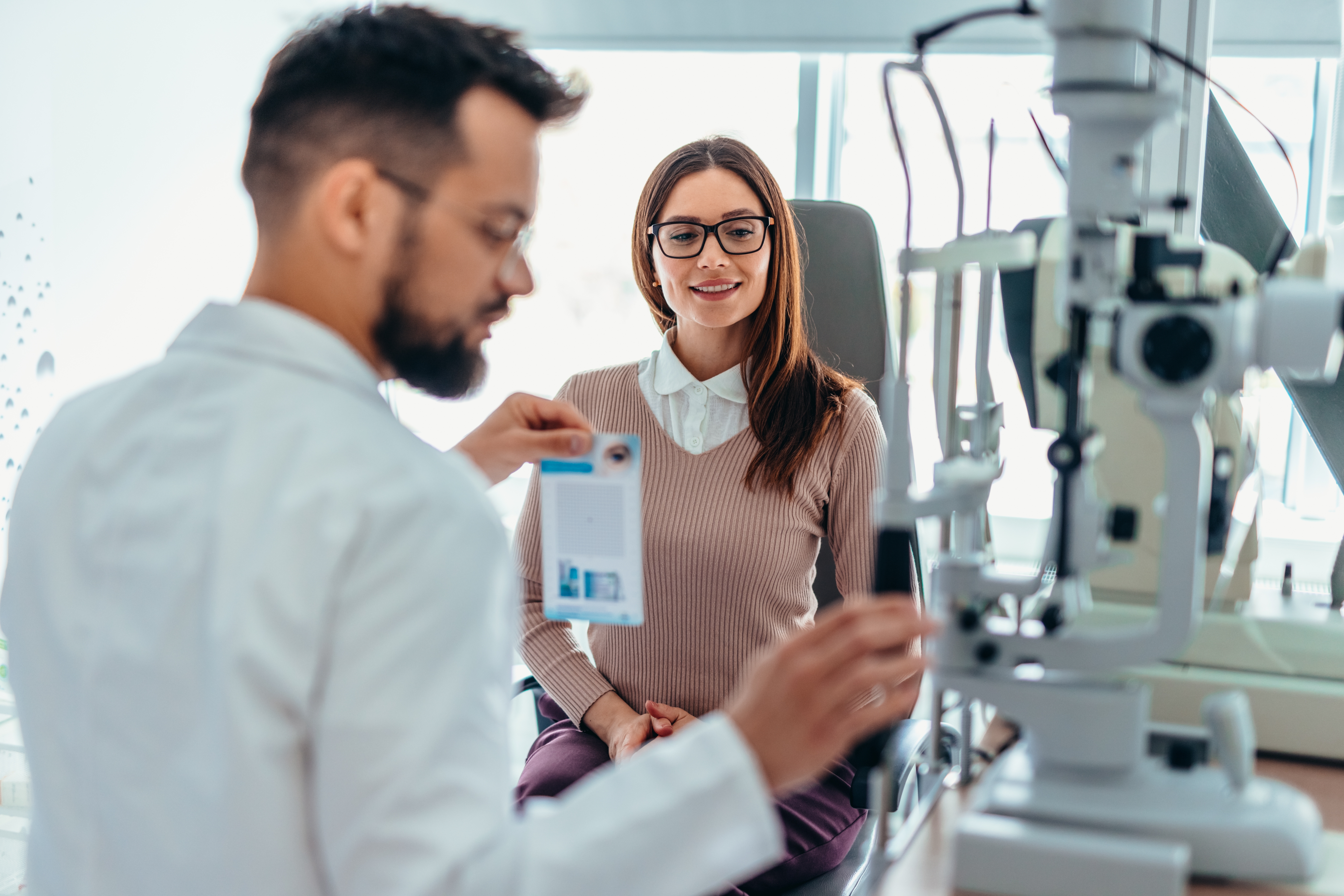 Doctor examines a woman with an eye test machine in an optometry office. The woman is seated and smiling, wearing glasses and business casual attire