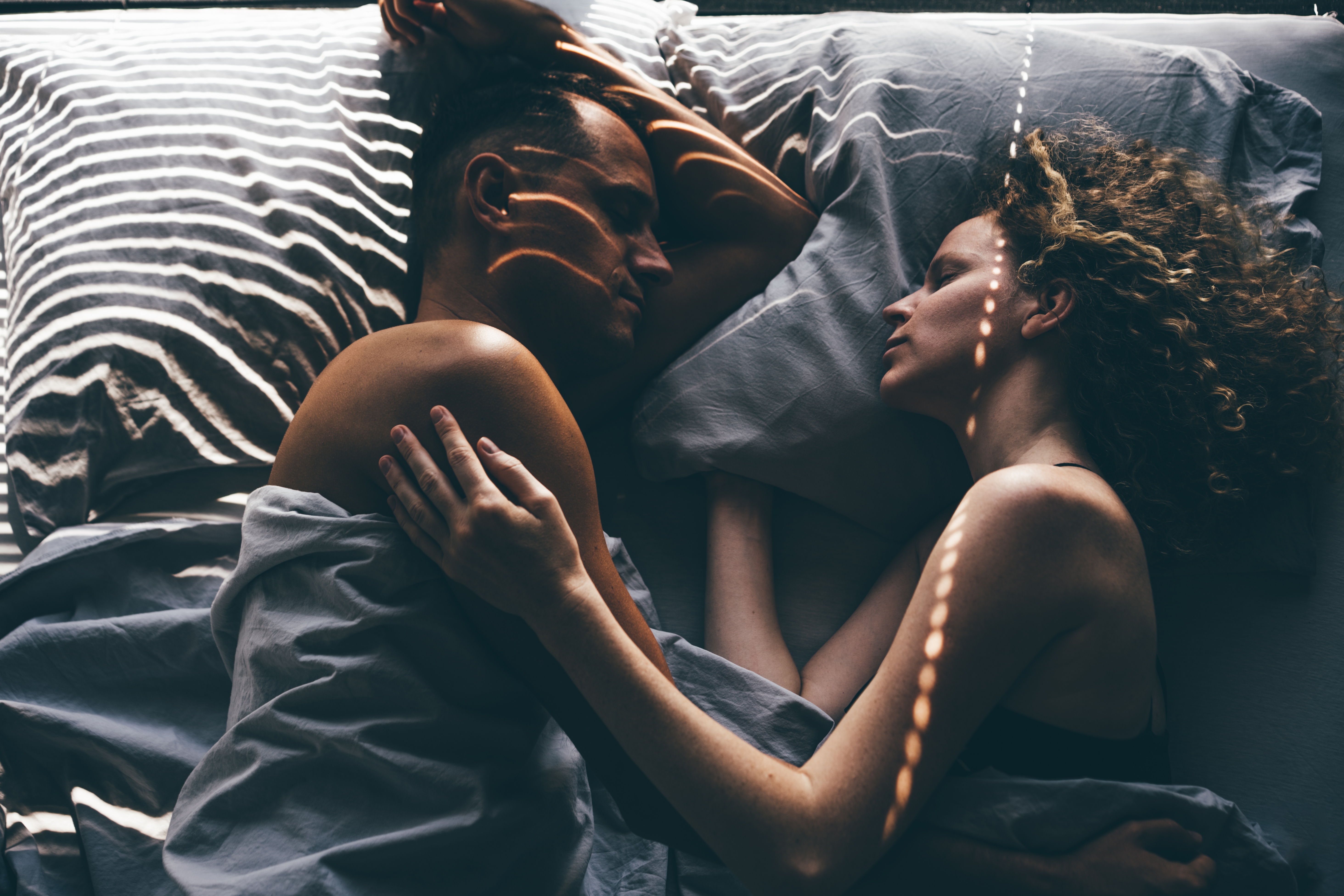 Two people lying in bed facing each other, wrapped in sheets, with an intimate and tender expression, light filtering through blinds creating patterns on them