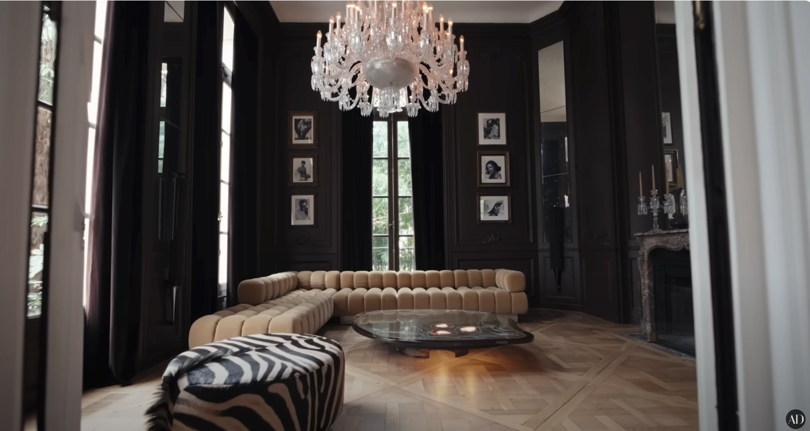 Elegant modern living room with large chandelier, sectional sofa, patterned ottoman, and floor-to-ceiling windows framed by dark curtains
