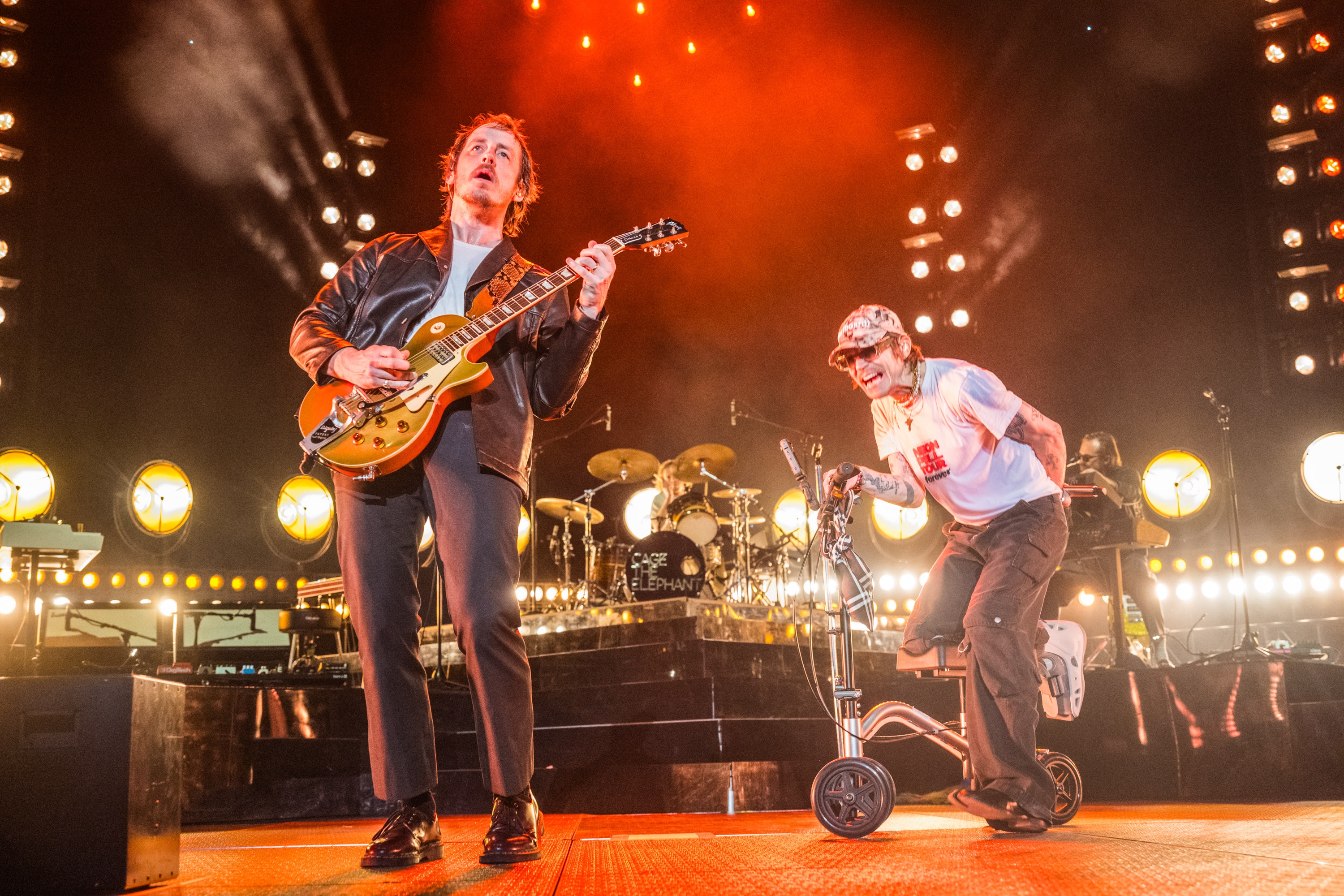 Two musicians performing energetically on stage; one with a guitar, the other using a knee scooter. Bright stage lights illuminate the scene