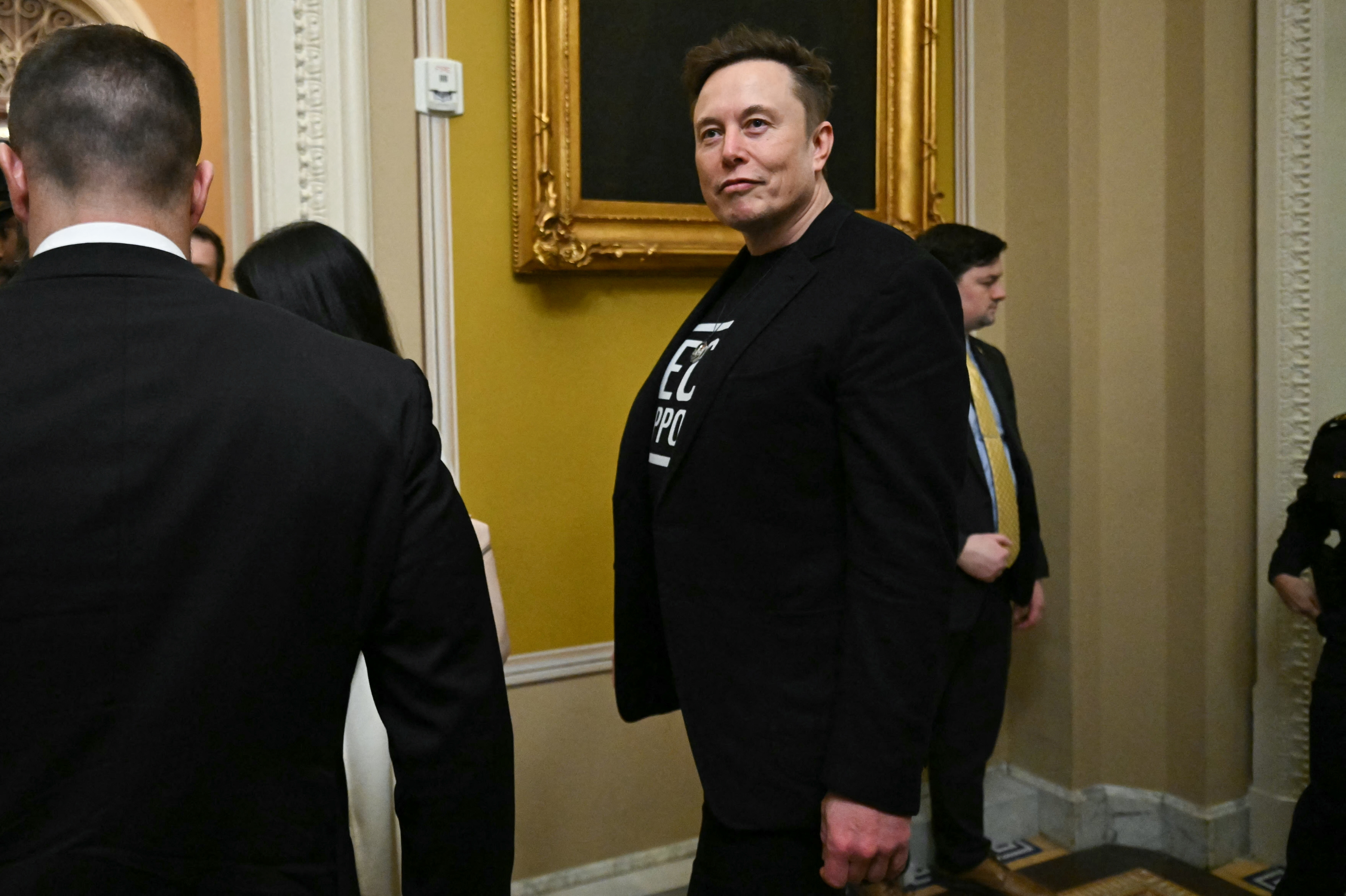A man in a suit and graphic t-shirt stands in a formal setting with others nearby, possibly at an event. A painting and ornate decorations are visible