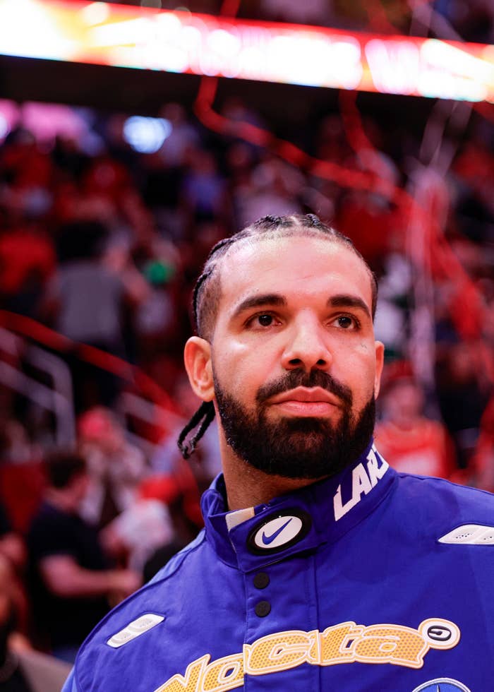 Drake with braided hair wearing a blue sports jacket looks on in a busy indoor arena setting