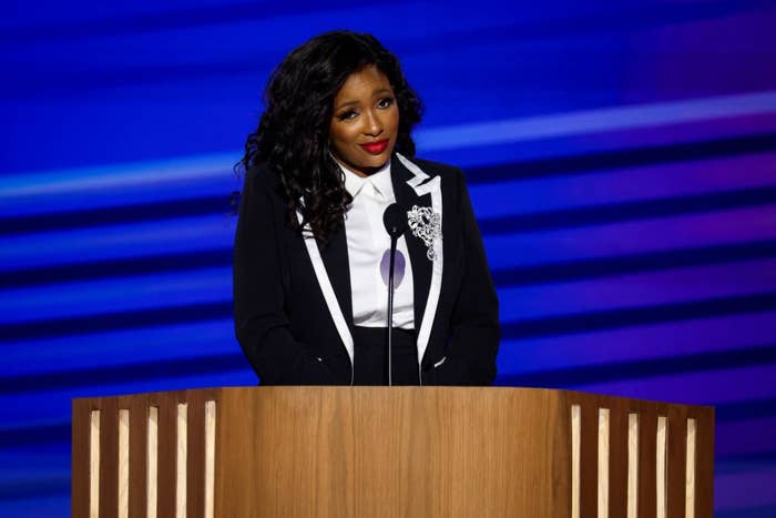 Person at a podium speaking, wearing a formal suit with a white shirt and decorative lapel detail