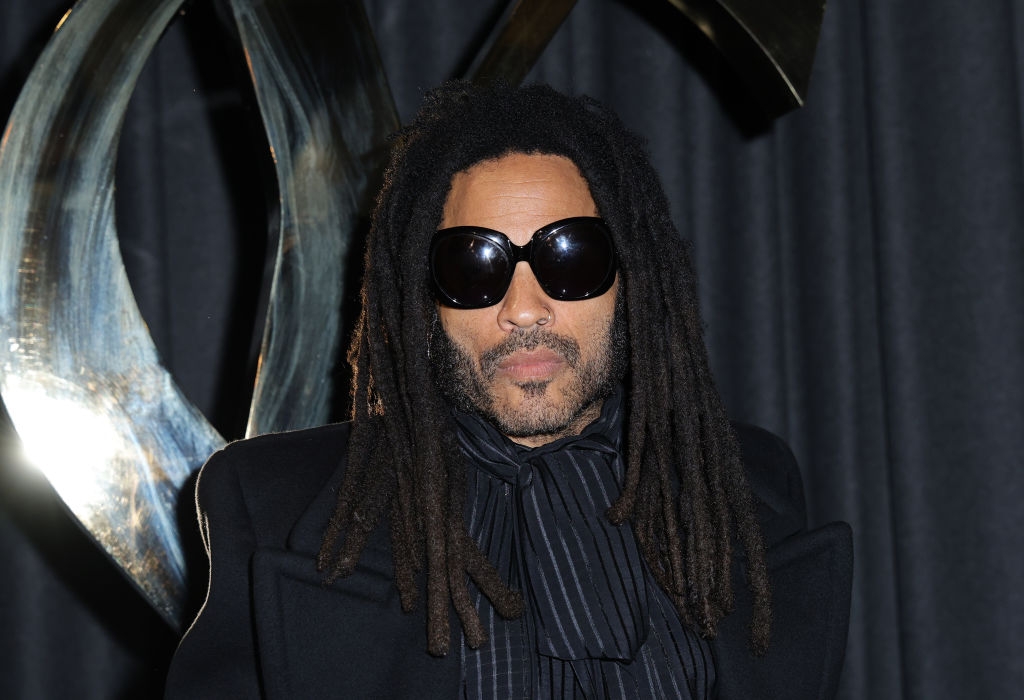 Person with dreadlocks wearing large sunglasses, a dark jacket, and a striped scarf at a formal event