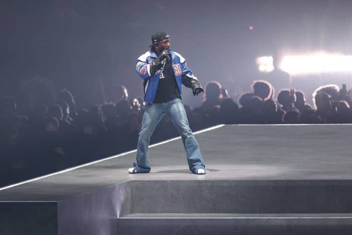 Kendrick Lamar in a sporty jacket and jeans stands on a stage, microphone in hand, engaging with a concert audience