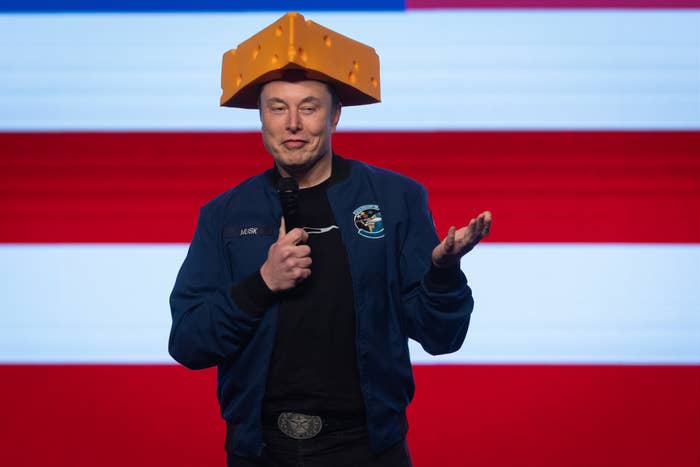 A person in a jacket holds a microphone, wearing a large cheese wedge hat, in front of an American flag background