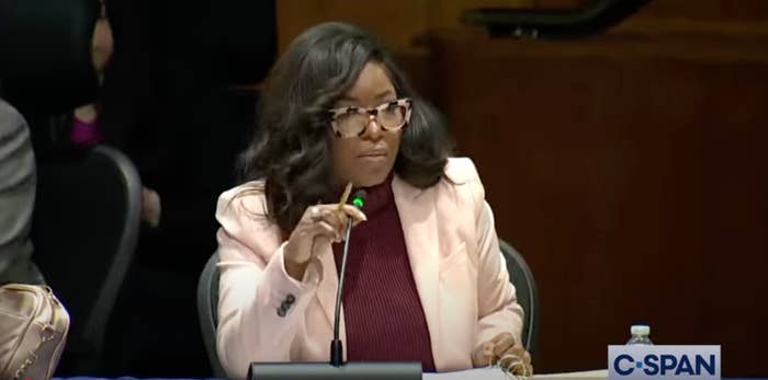 Person speaking at a microphone during a formal meeting, wearing glasses and a stylish blazer over a turtleneck