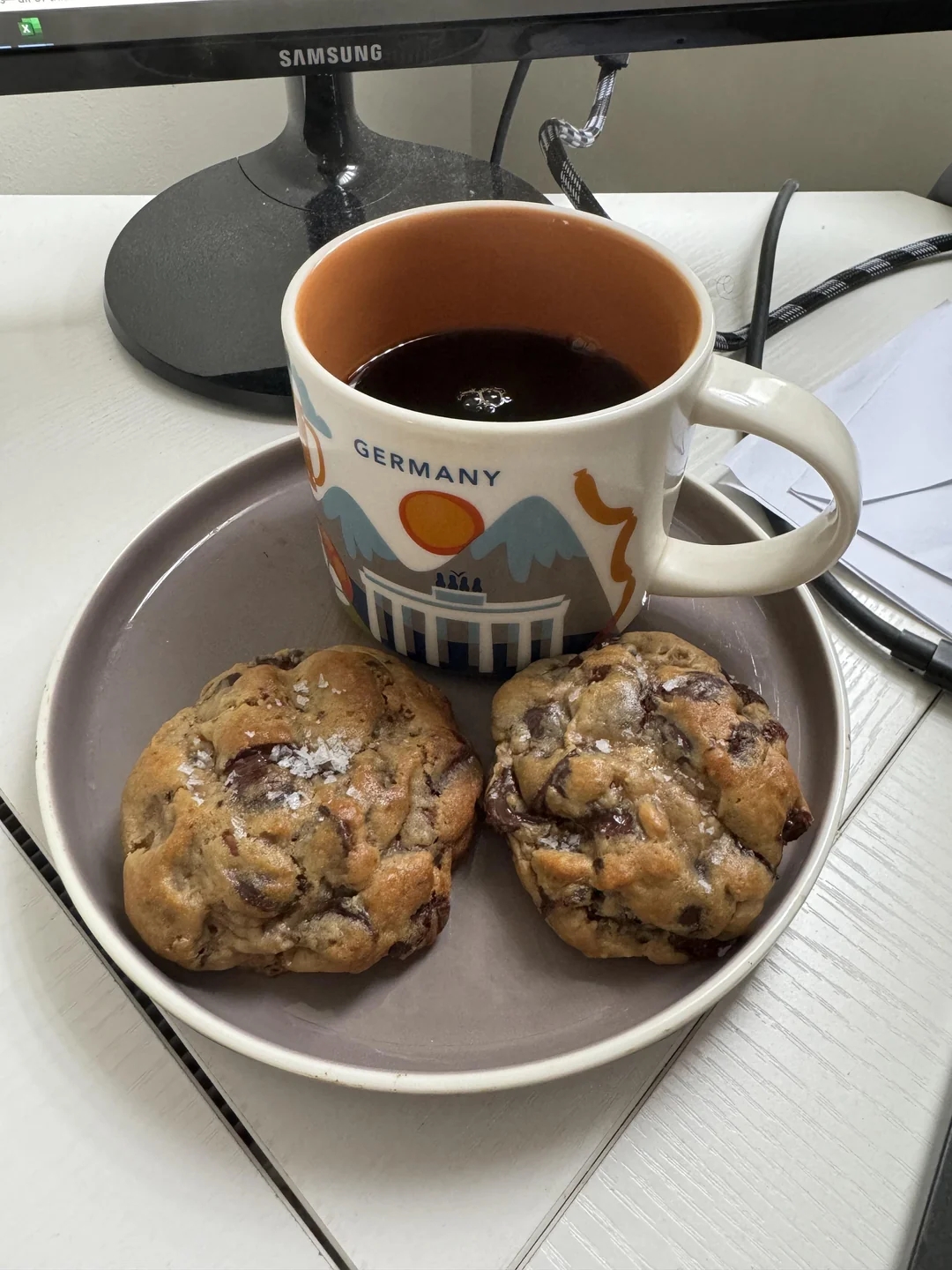 A desk with a coffee in a "Germany" mug and two chocolate chip cookies on a plate, next to papers and a computer monitor