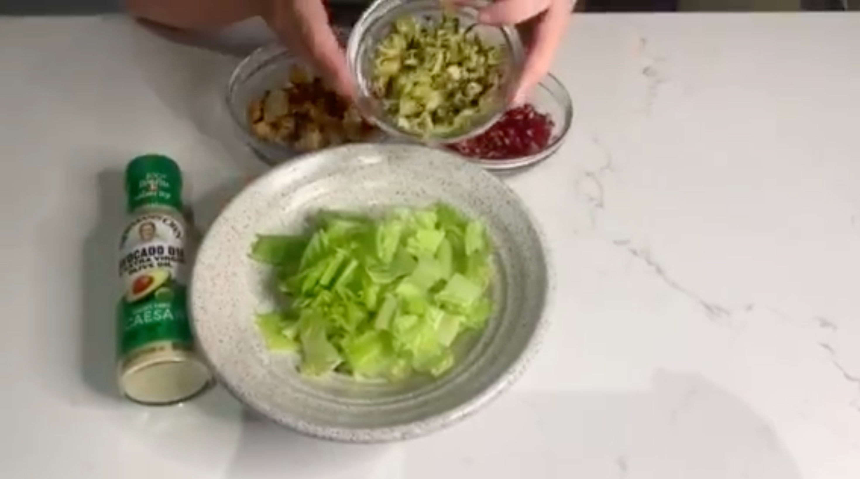 Person preparing a salad with chopped lettuce, croutons, diced avocado, and pomegranate seeds on a countertop. Avocado oil spray nearby