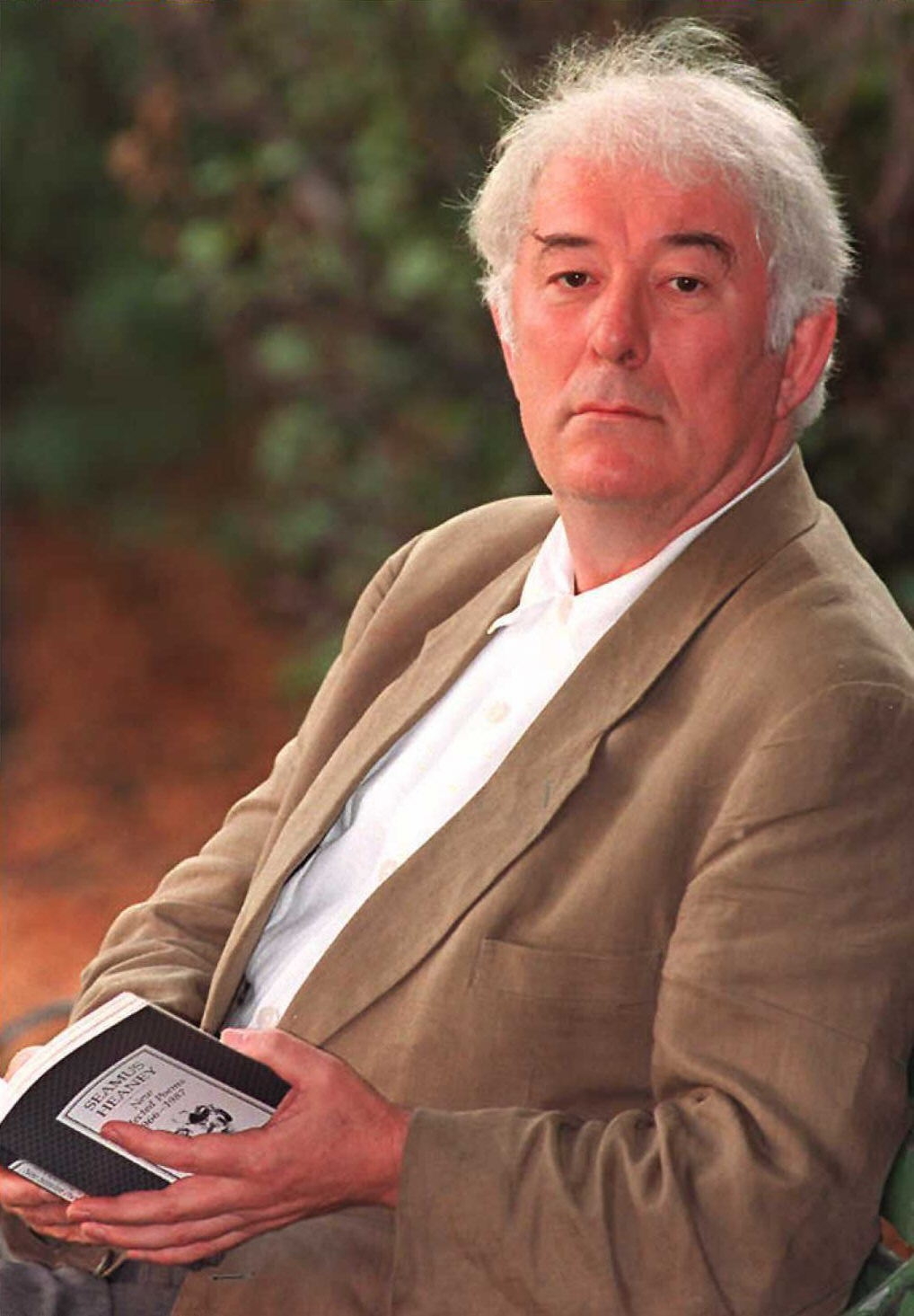 A person sits outdoors holding a book, wearing a light blazer and shirt, with a contemplative expression