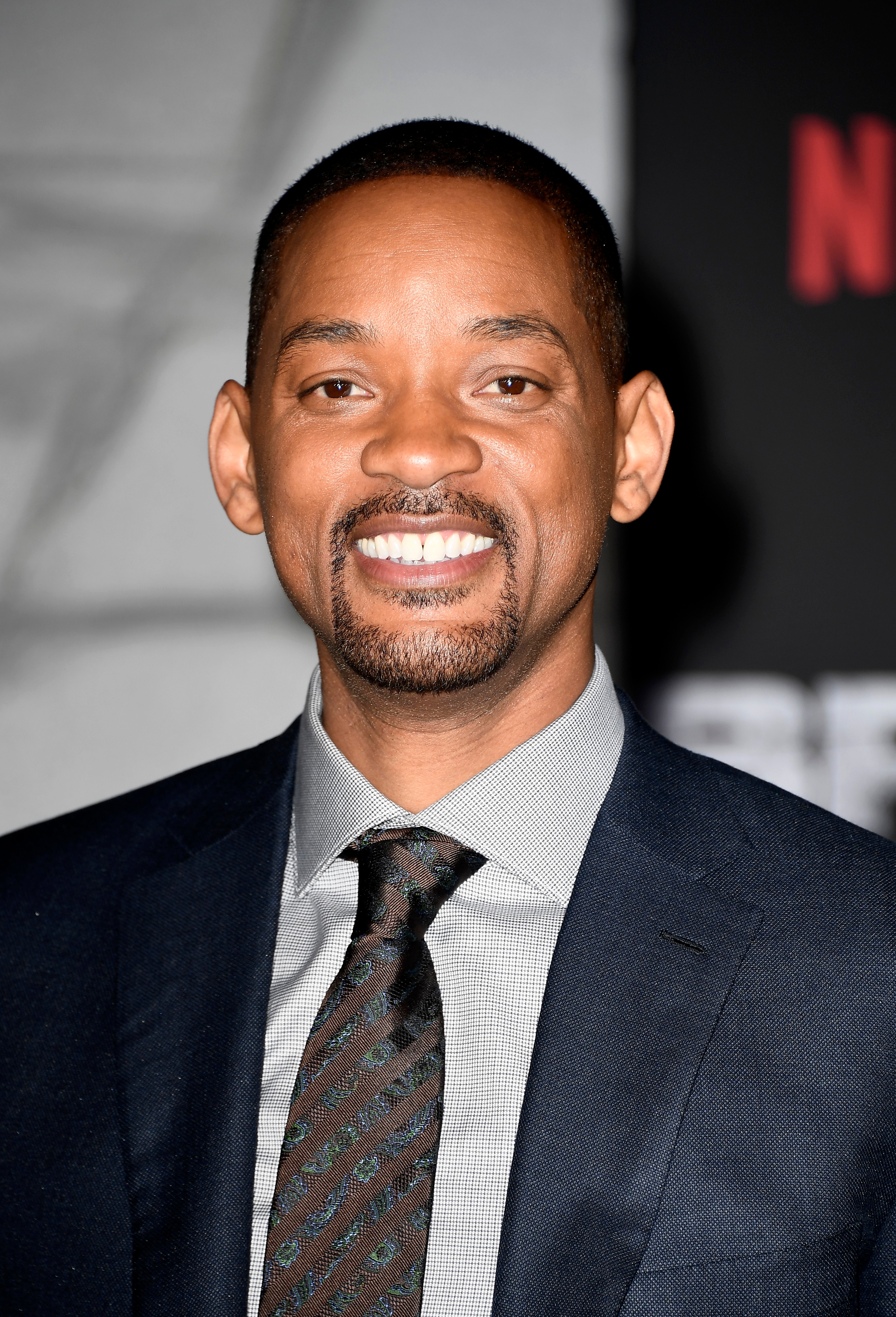 A person in a formal suit smiles at a media event, standing in front of a Netflix backdrop