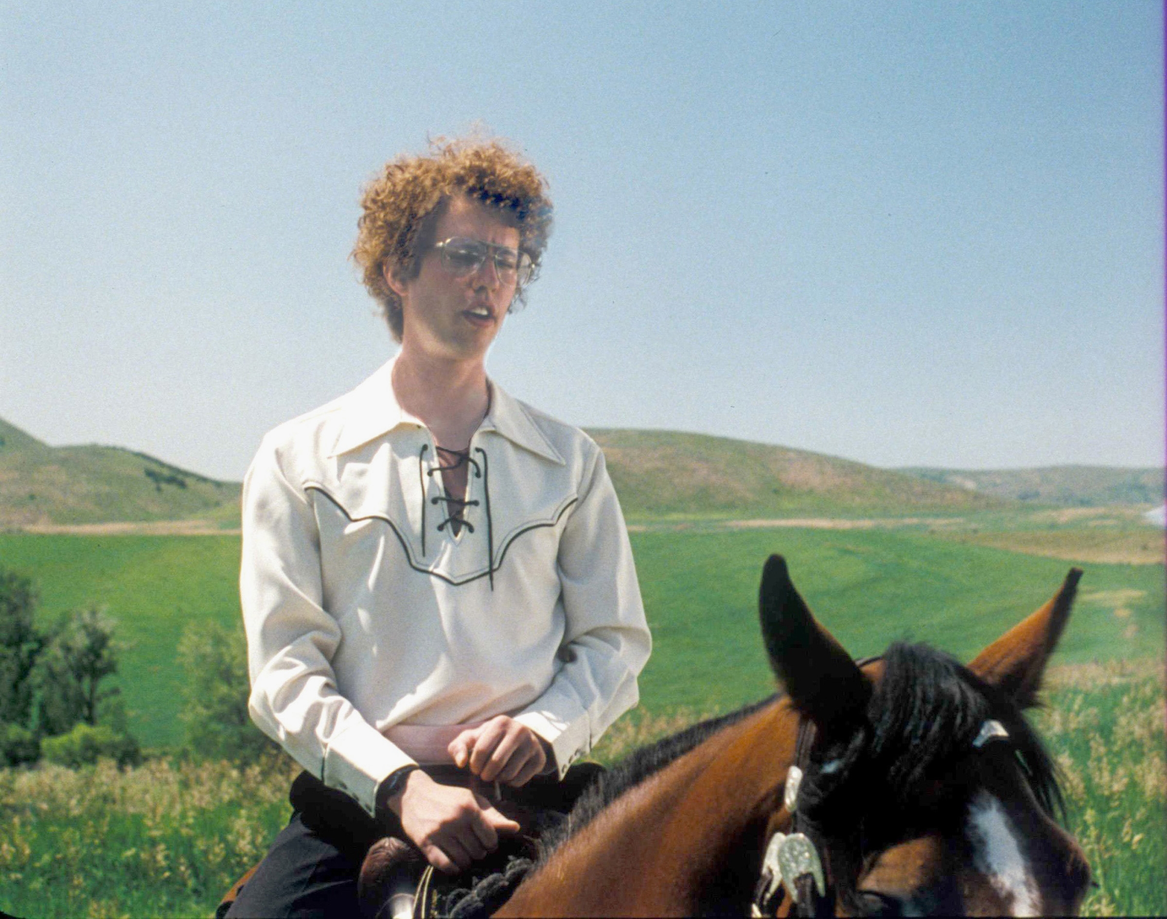 Person with curly hair, wearing a vintage-style shirt with embroidery, seated on a horse with a rural landscape in the background