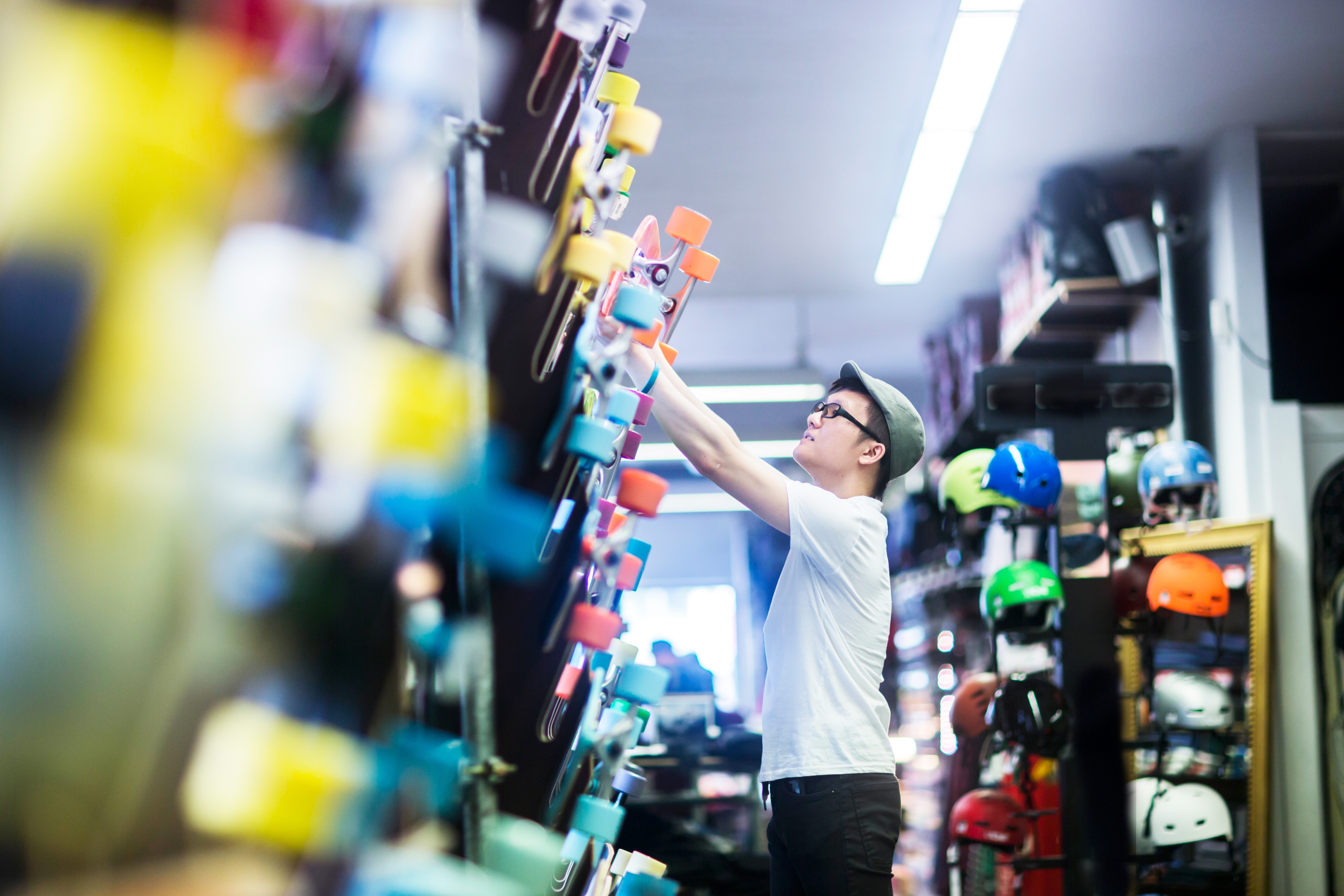 Person in a hat and glasses reaches for a skateboard in a shop filled with various sports gear and accessories