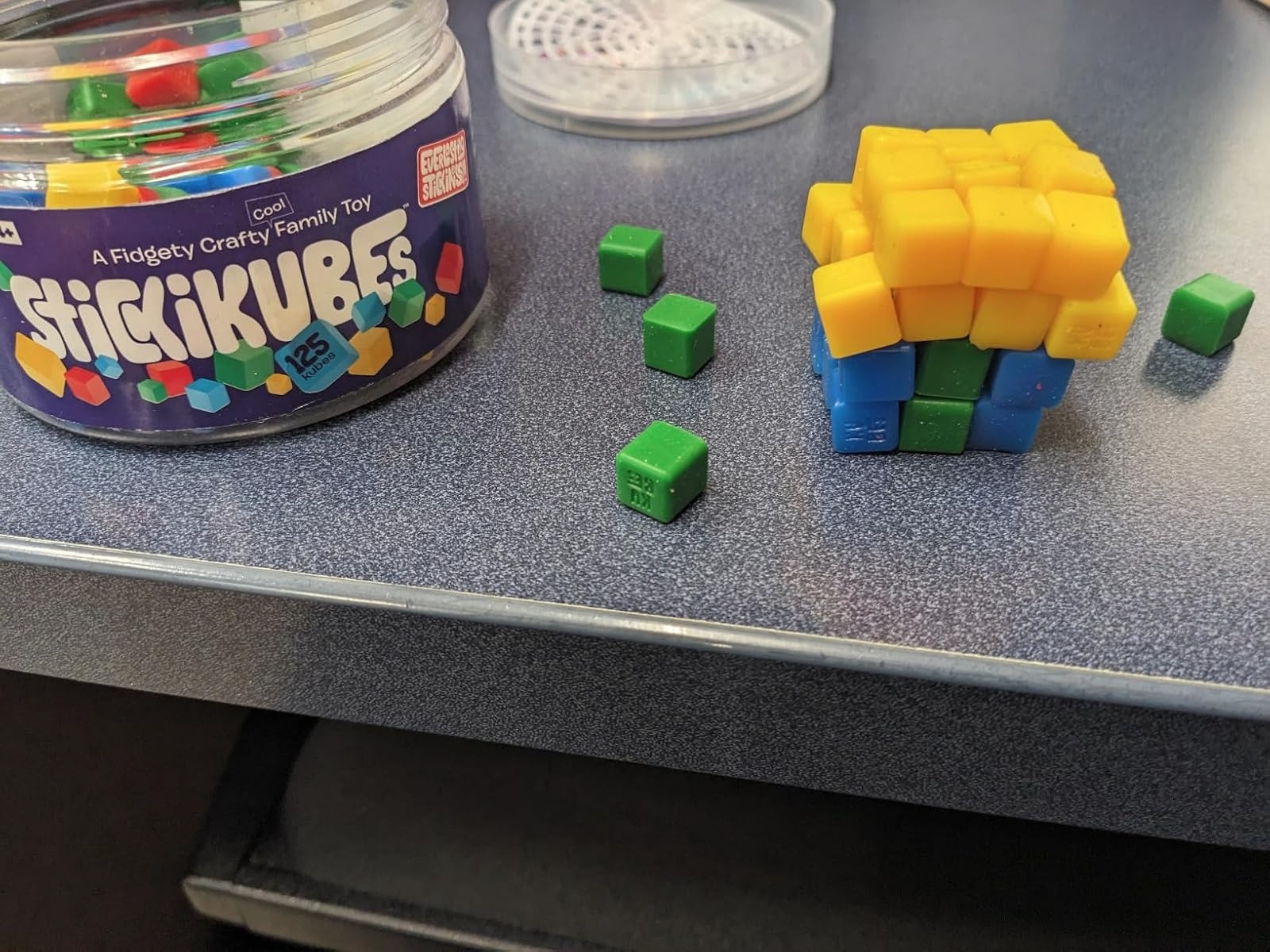 StickiKubes toy jar beside partially assembled cube made of green and yellow pieces on a desk