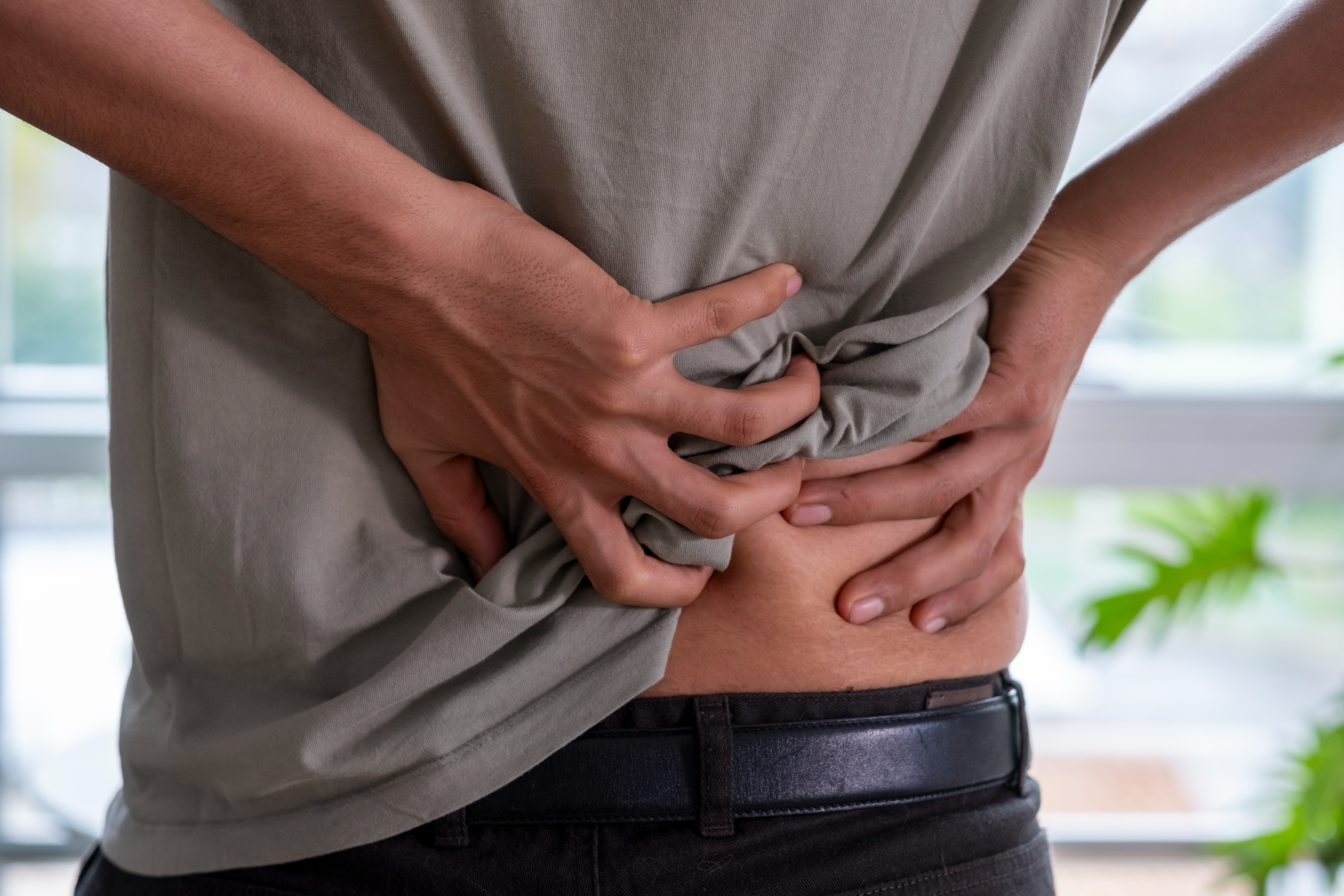 Person clutching their lower back in discomfort, suggesting back pain. Hands grip the shirt and waist area for support