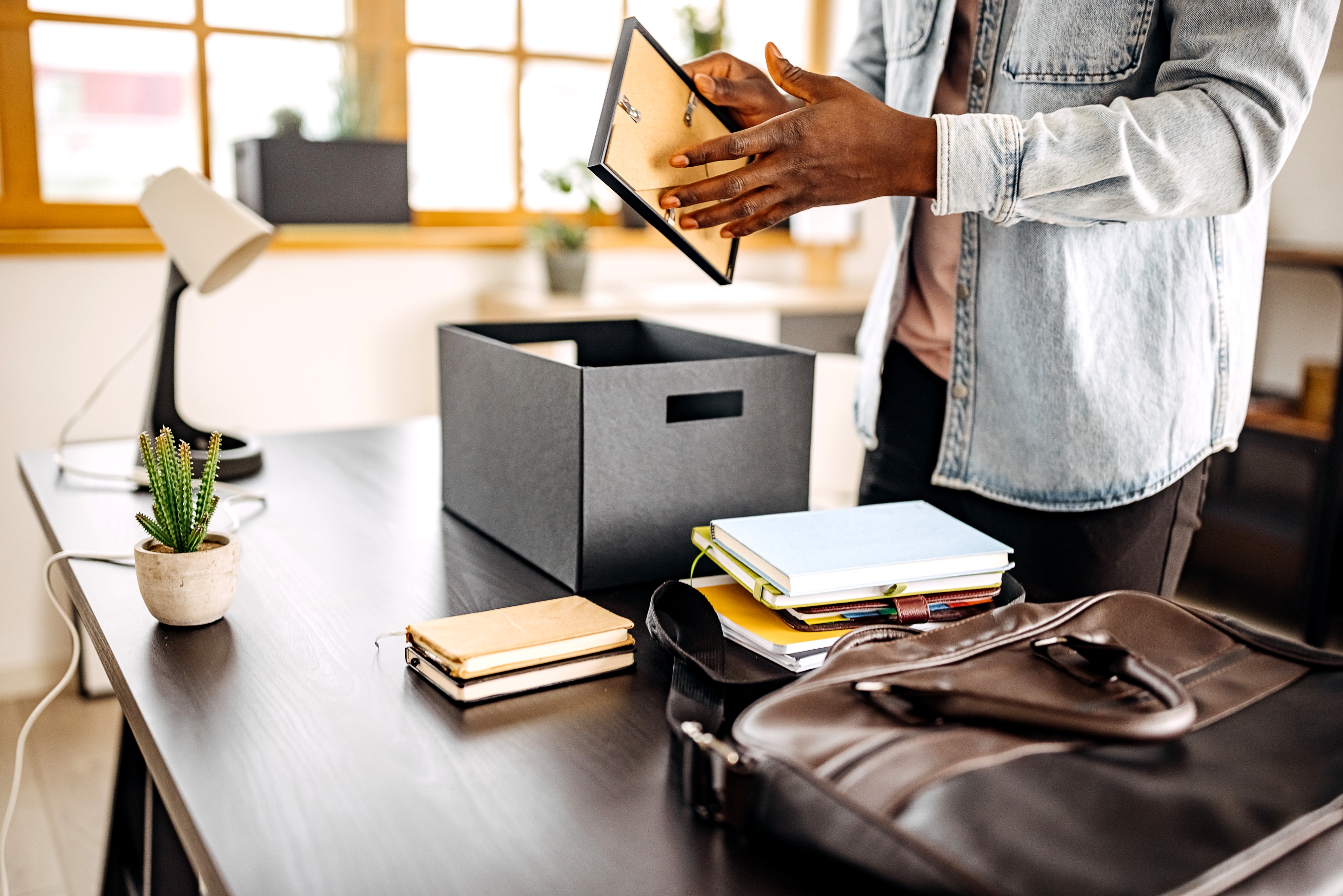 Person packing office items into a box, including files and a tablet, suggesting a job change or office move