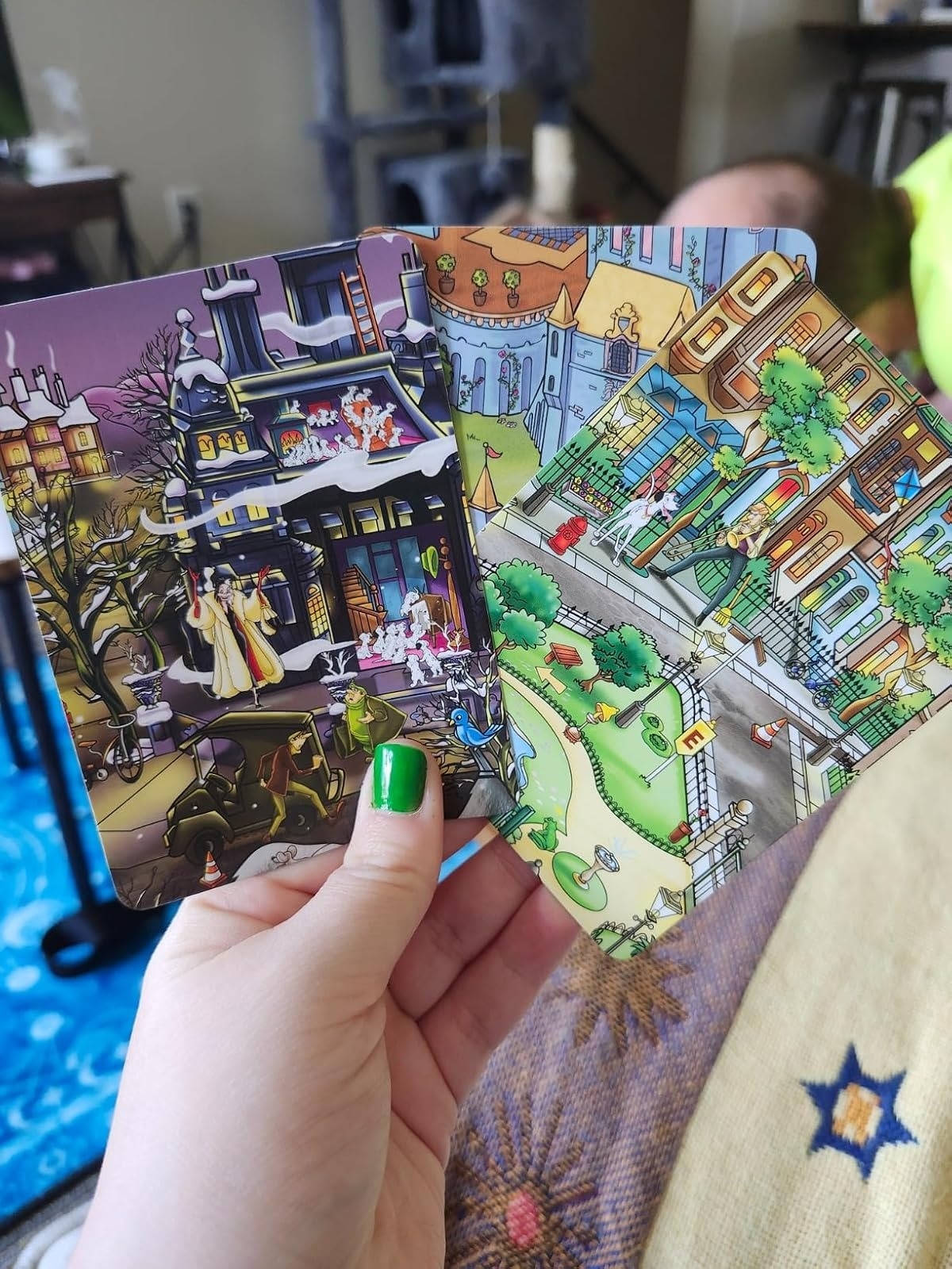 Person holding vibrant illustrated cards depicting winter, a castle, and a park. One fingernail is painted. Room background is slightly blurred