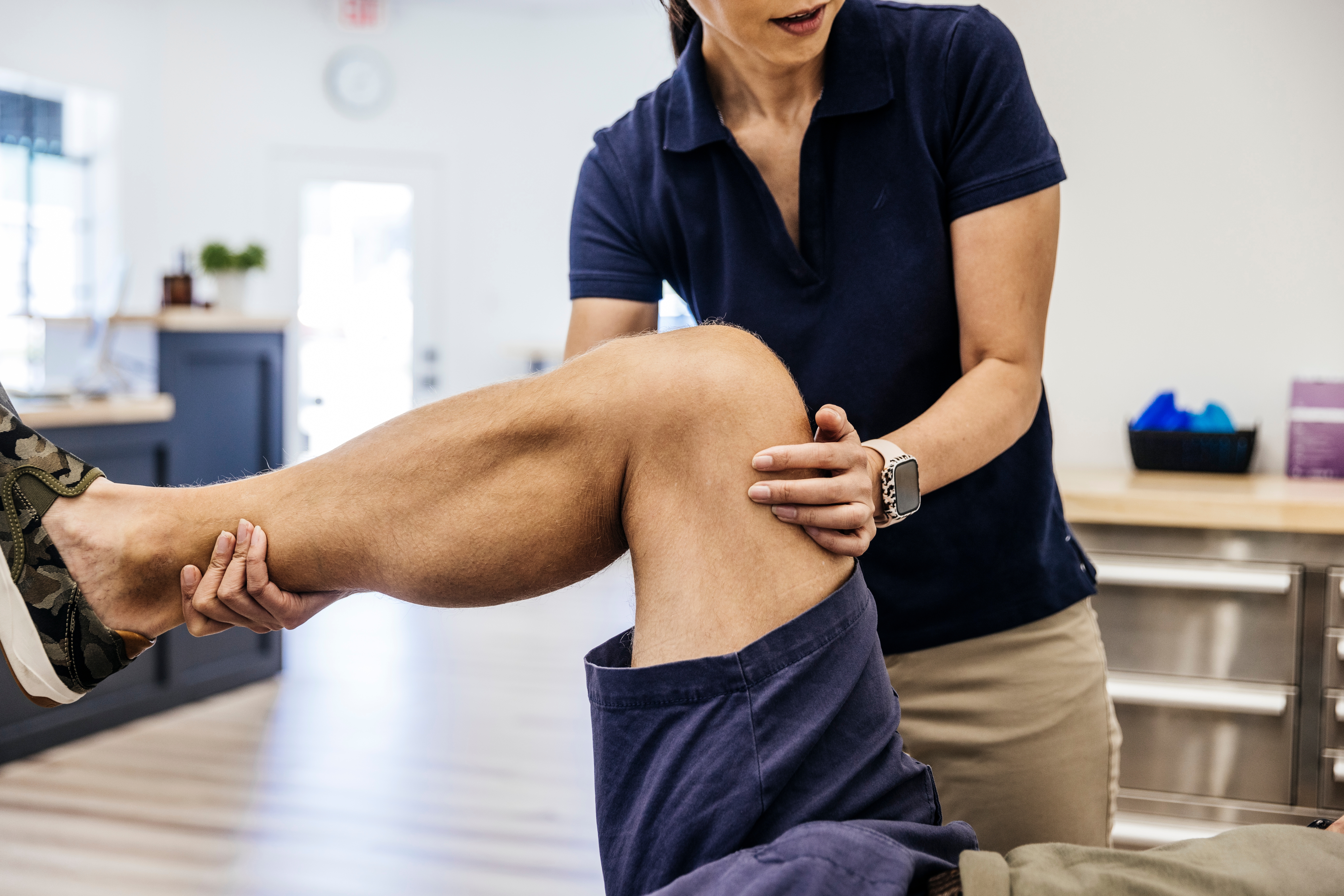 A physical therapist assists a patient by supporting their bent leg during a therapy session in a clinic setting