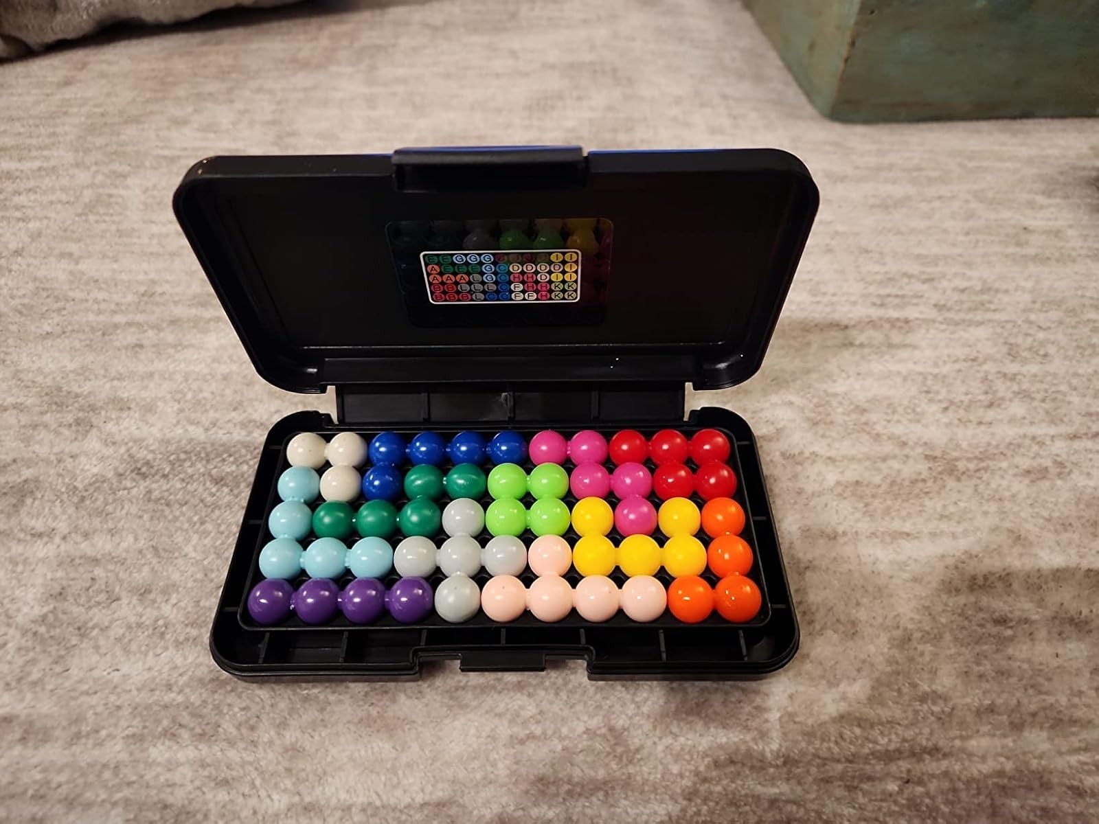 Open case with colorful, spherical beads arranged in a grid pattern, possibly a puzzle or game set, on a textured surface