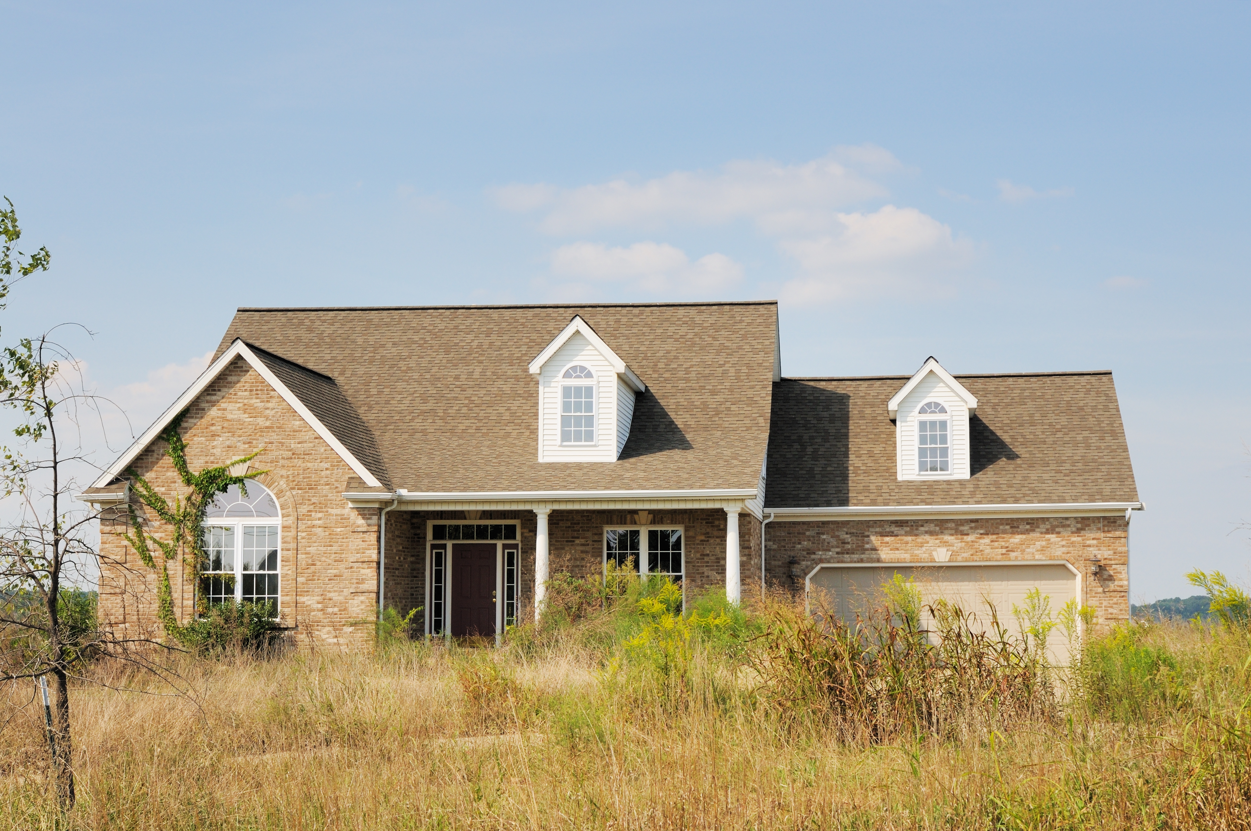 A modest brick suburban home with dormer windows and a two-car garage, surrounded by overgrown grass and vegetation, under a clear sky