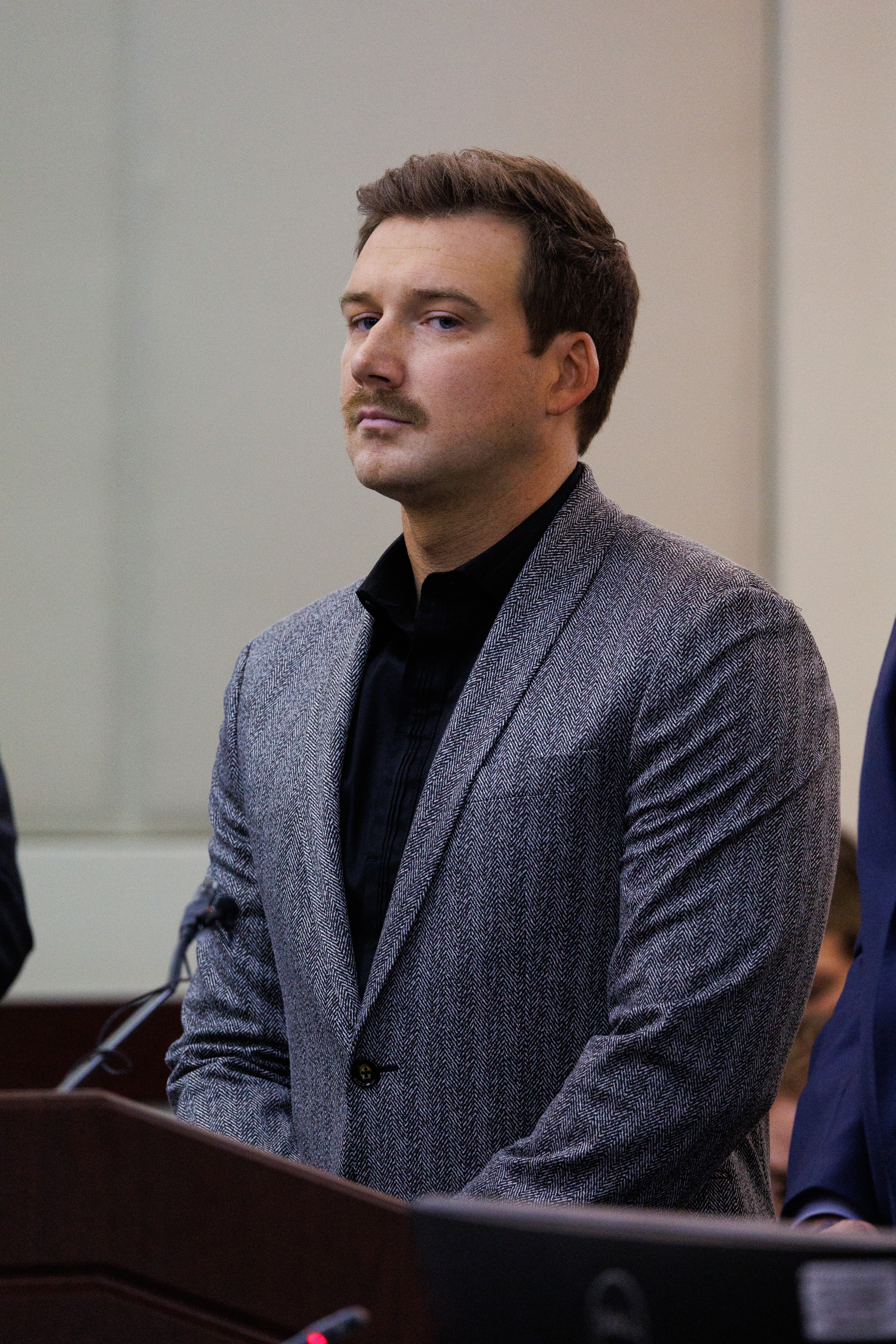 Man in a formal setting wearing a textured suit jacket and dark shirt, standing attentively behind a podium