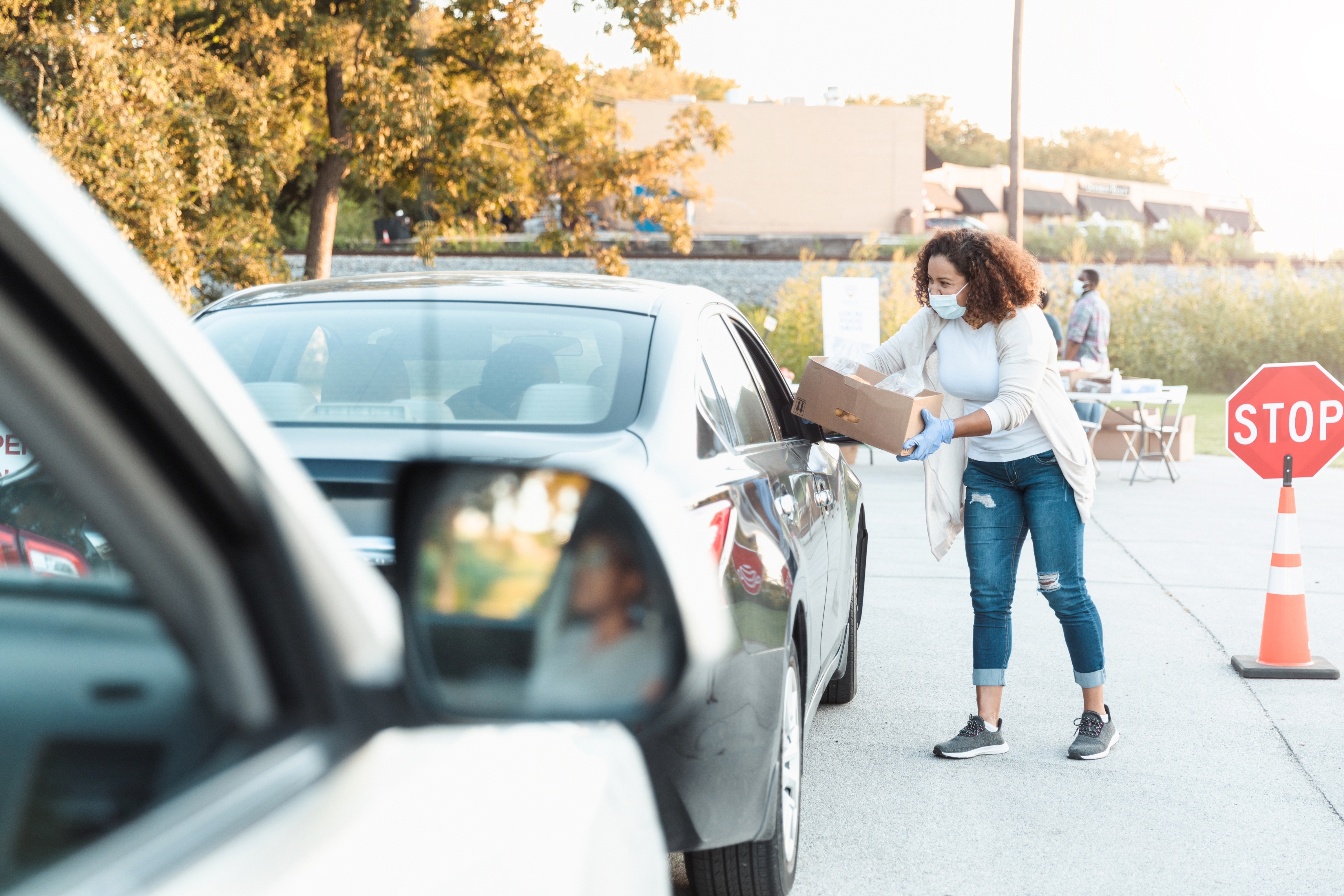 A woman in a mask and gloves hands a box to a driver through their car window at a roadside stop, highlighting safe work practices
