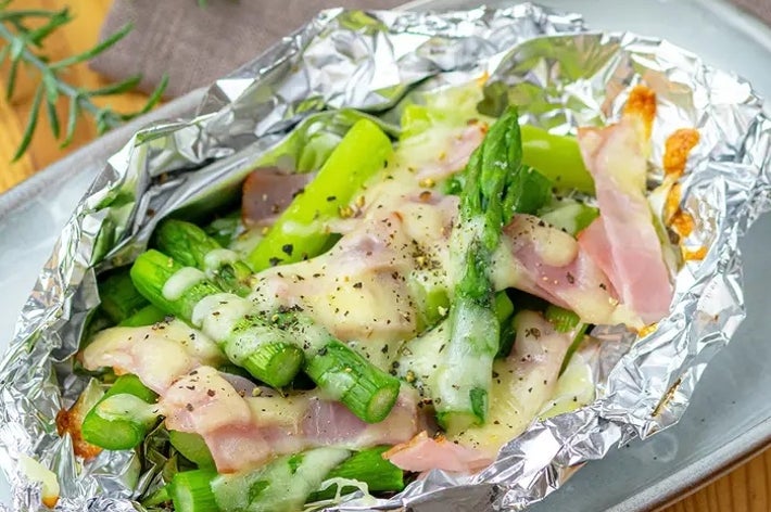 ホイルに包まれたアスパラガスとハムの料理。焼かれていてチーズがかかっている。