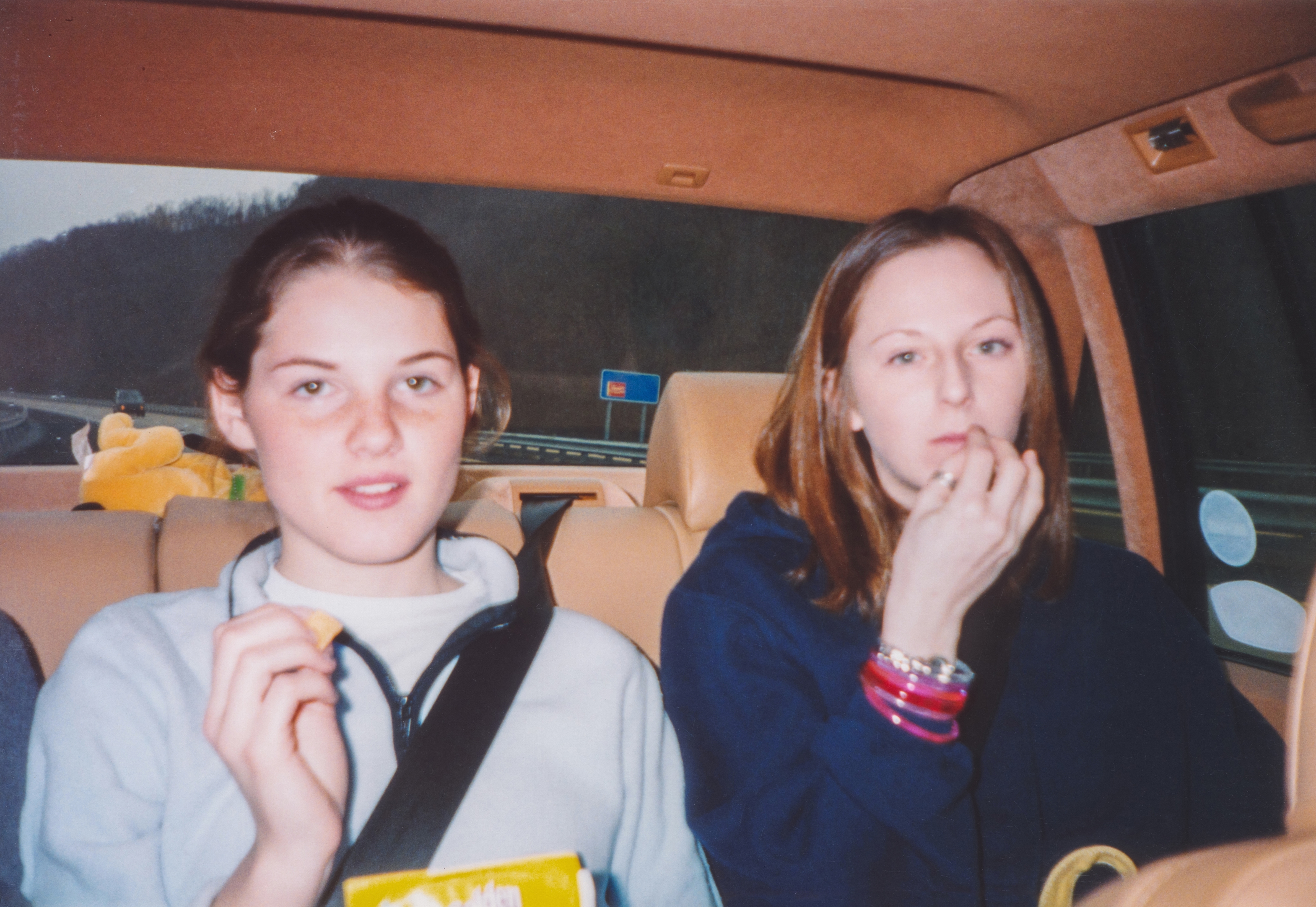 Two people seated in a car, each holding snacks. The person on the left holds a box, and the person on the right has a stack of colorful bangles