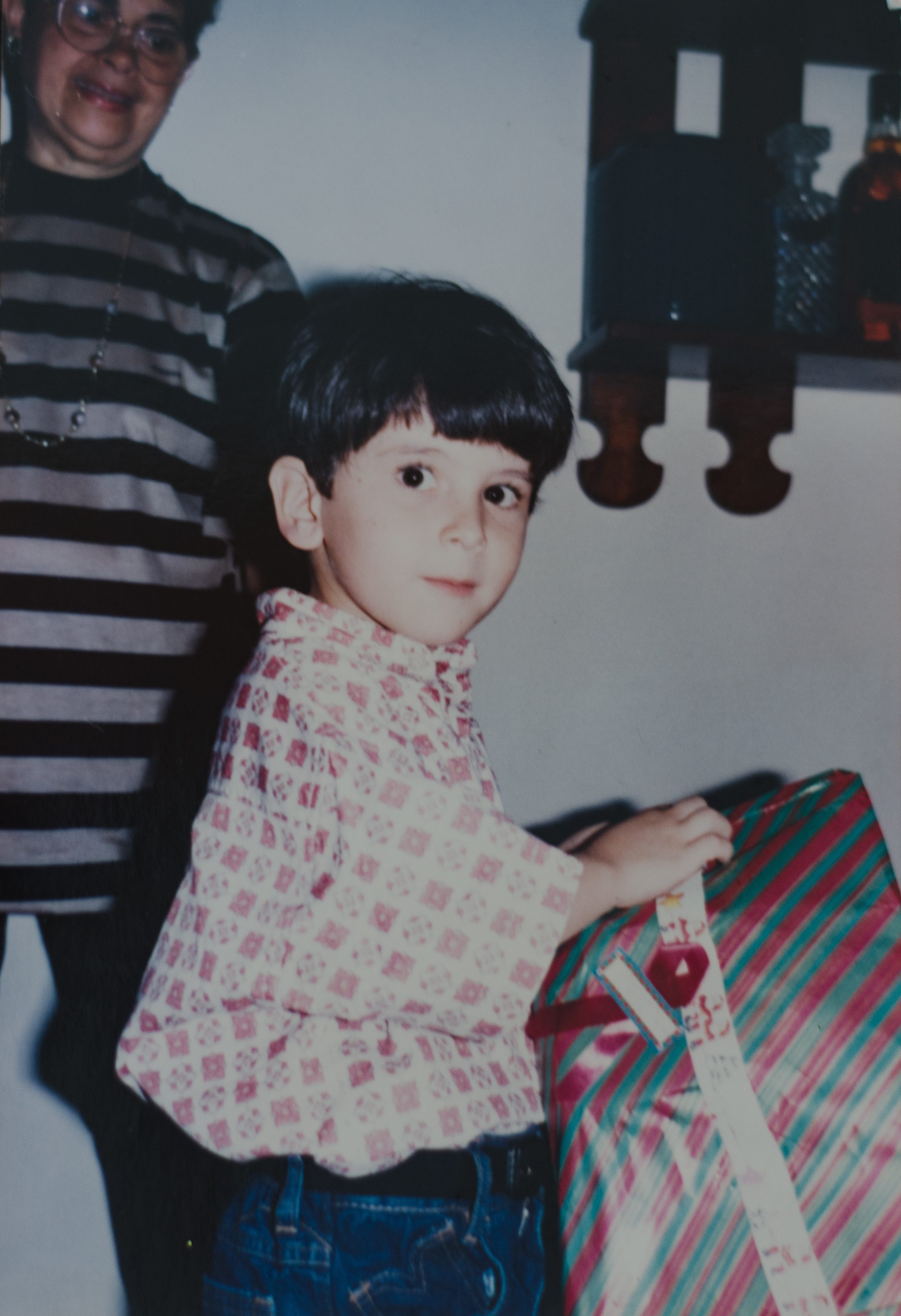Child holding a gift with striped wrapping, standing next to an adult. The child wears a patterned shirt and jeans, looking towards the camera