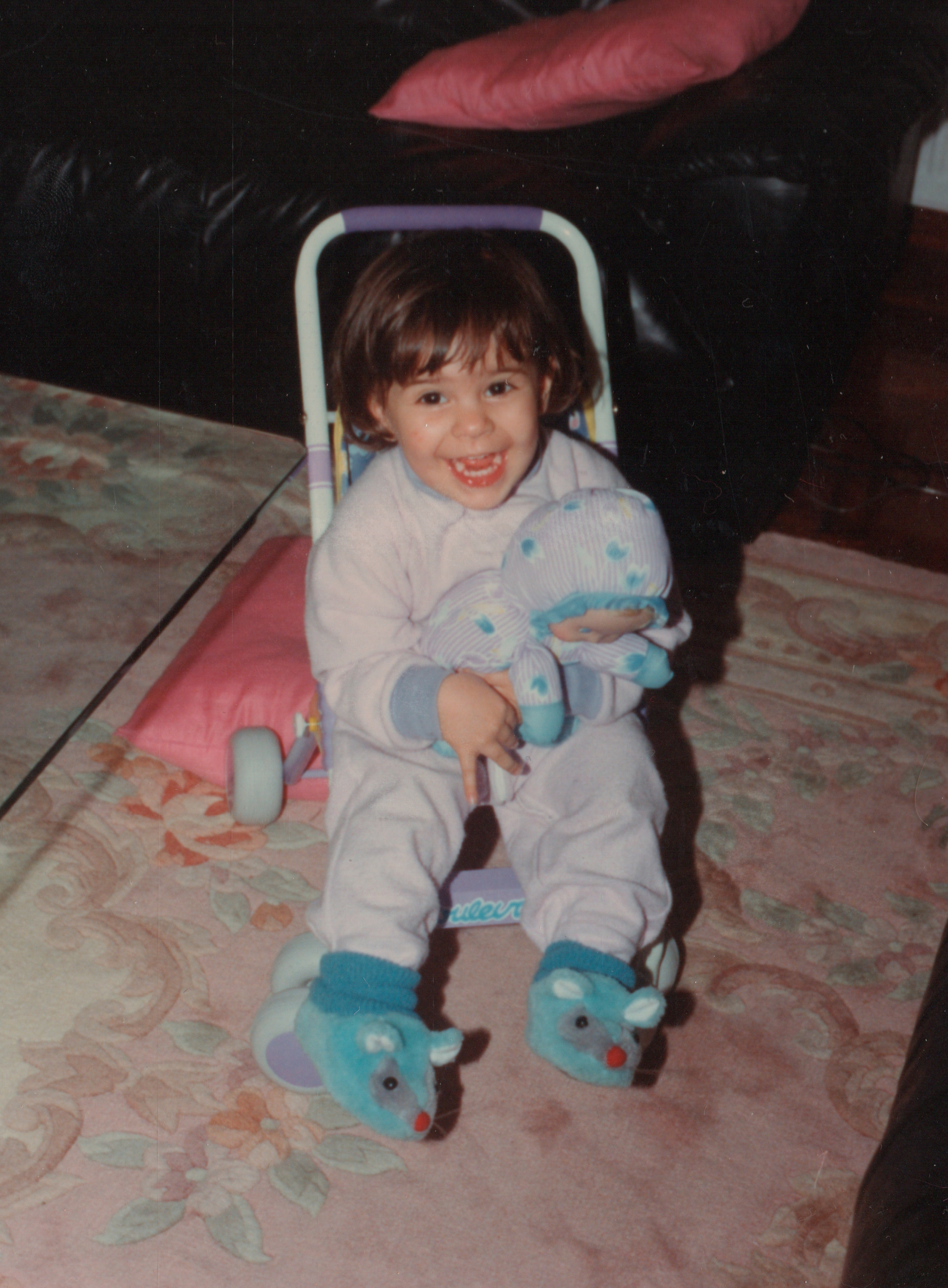 A child smiling while sitting on a toy stroller, holding a stuffed animal, wearing pajamas and animal-themed slippers, in a cozy room