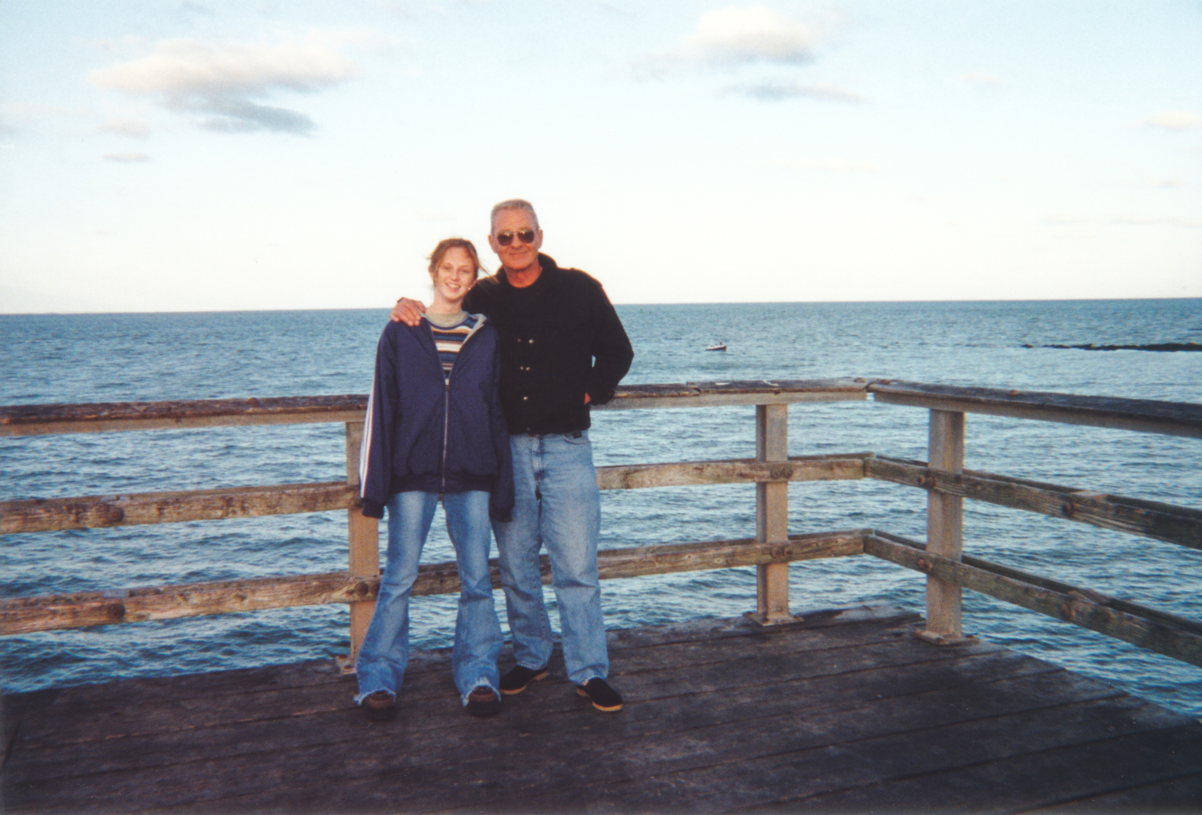Two people standing on a wooden pier by the ocean, smiling at the camera. The scene suggests a relaxed and casual outdoor moment